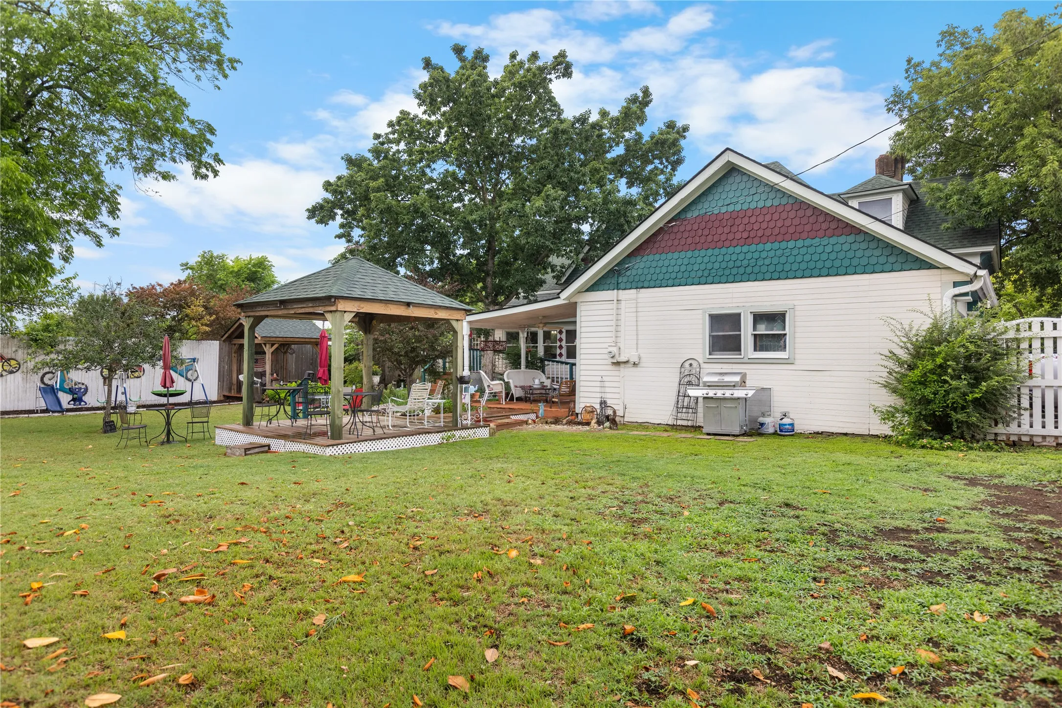 Spacious backyard featuring a built-in gazebo with a shingled roof, a raised wooden deck, a white picket fence, and a detailed gable facade with patterned shingle siding