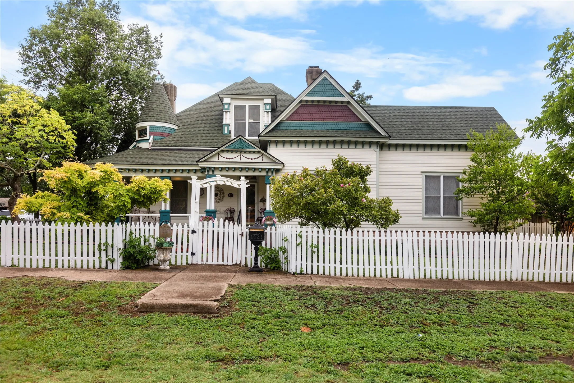 Victorian-style residence featuring a prominent turret, white picket fence, covered front porch with decorative trim, horizontal siding, and multiple gables with accent colors