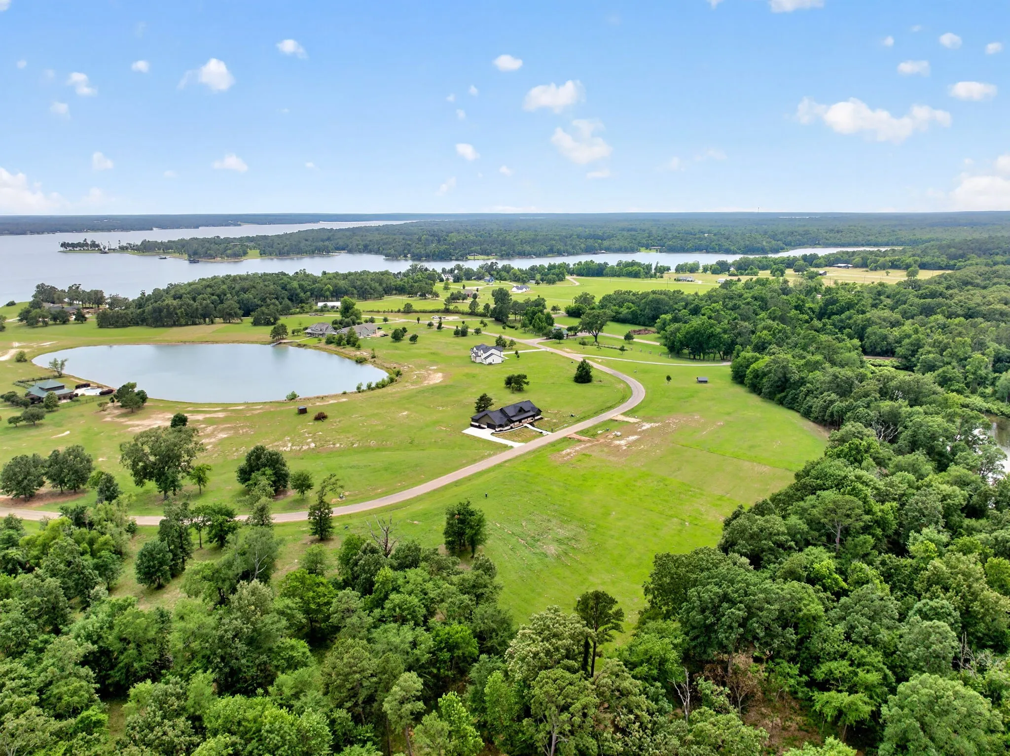 Expansive property featuring a tranquil lake, lush green fields, mature trees, and a winding driveway