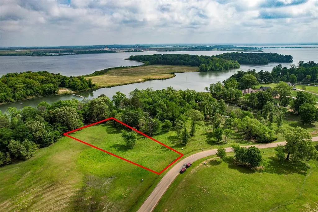 Expansive green lot bordered by mature trees and a paved road, featuring water frontage on a serene inlet