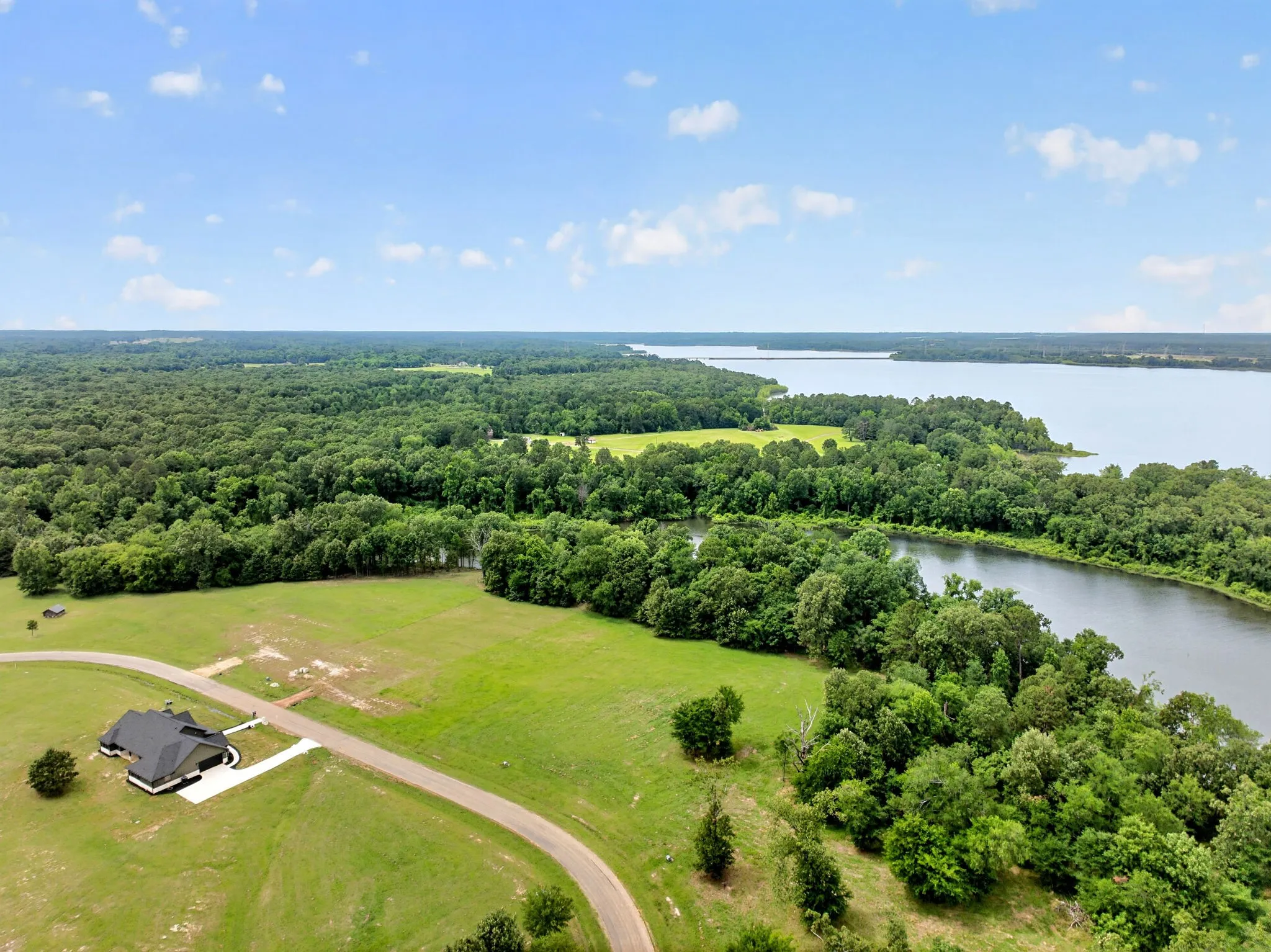 Expansive property featuring a single-story residence with a dark roof and light exterior, surrounded by green fields and dense woodland