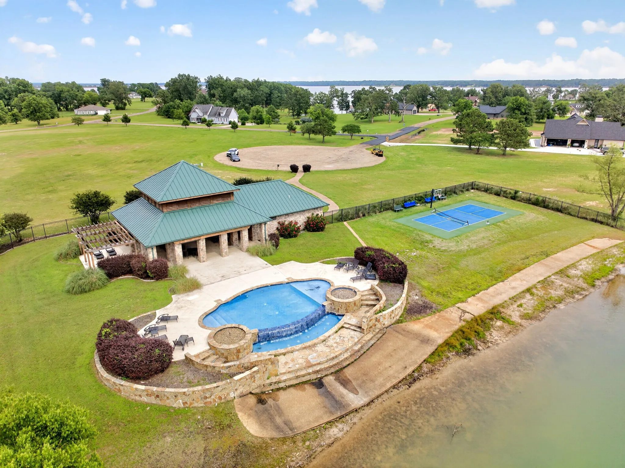 Community recreation area featuring a multi-level pool with stone surround, a clubhouse with a metal roof, and a fenced sports court