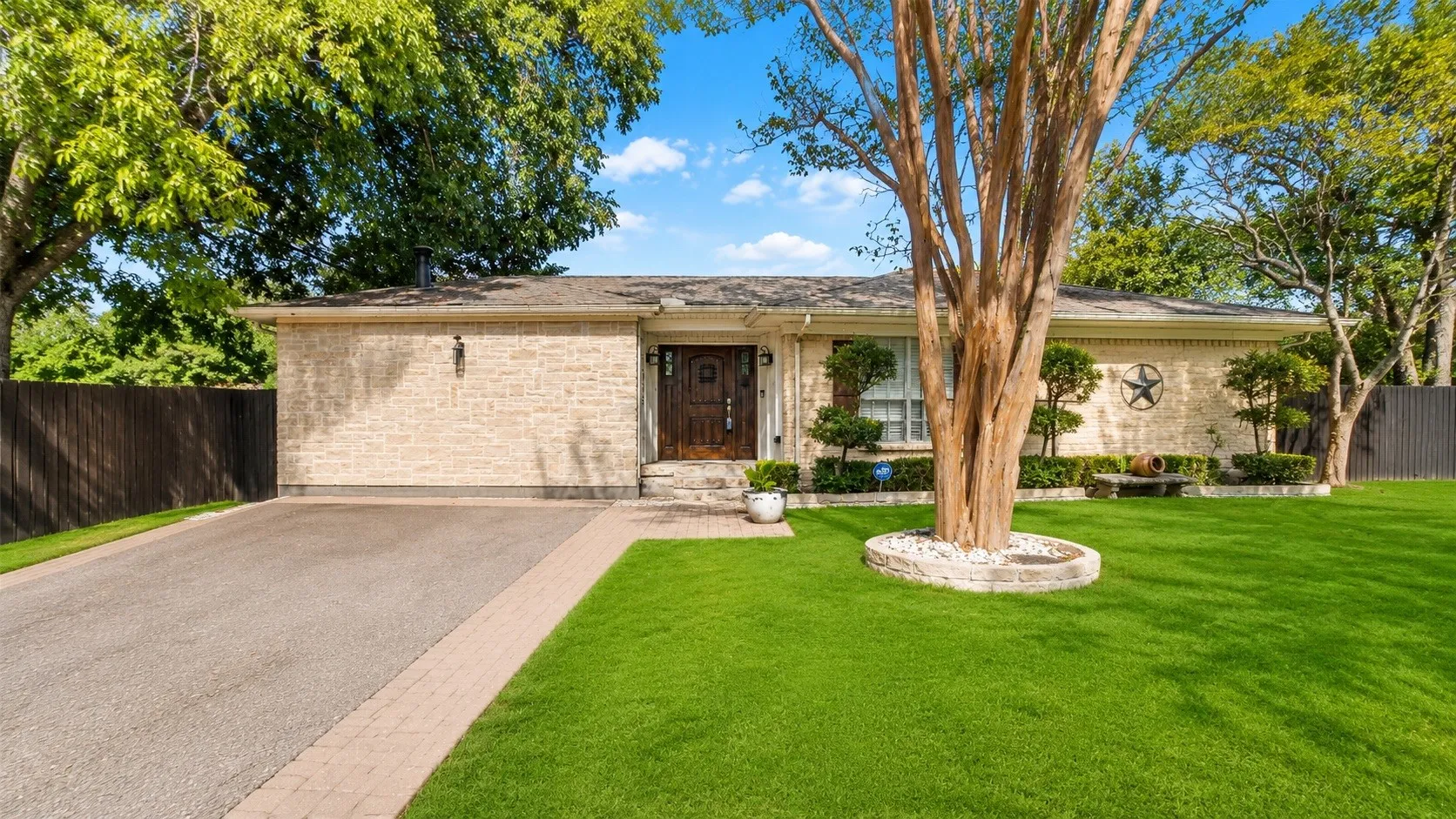 Stone facade home featuring a prominent front lawn, mature trees, a paved driveway with brick paver edging, and a dark wooden privacy fence