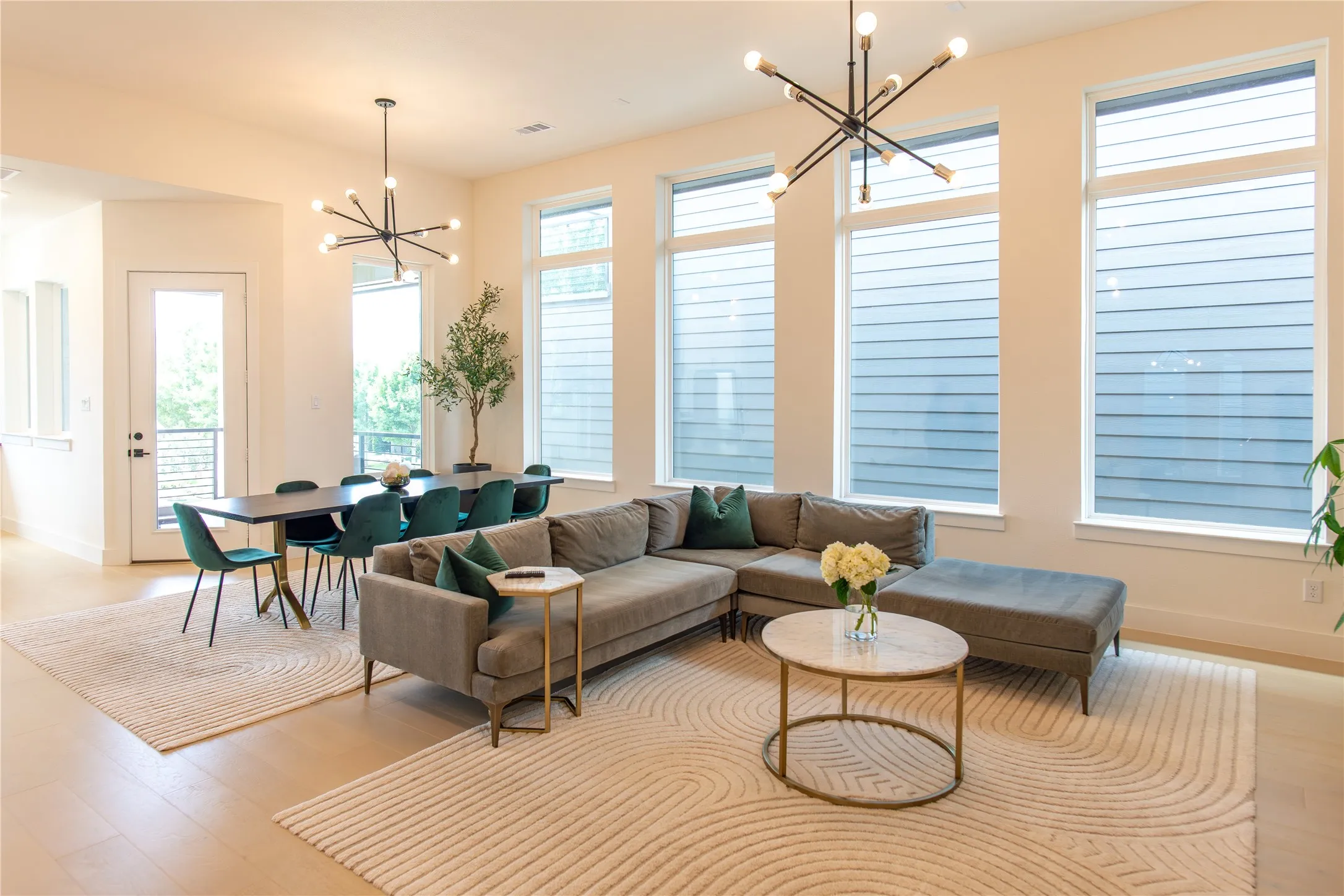 Open-concept living space with light-tone wood-finish flooring, high ceilings, multiple tall windows, and contemporary linear chandeliers
