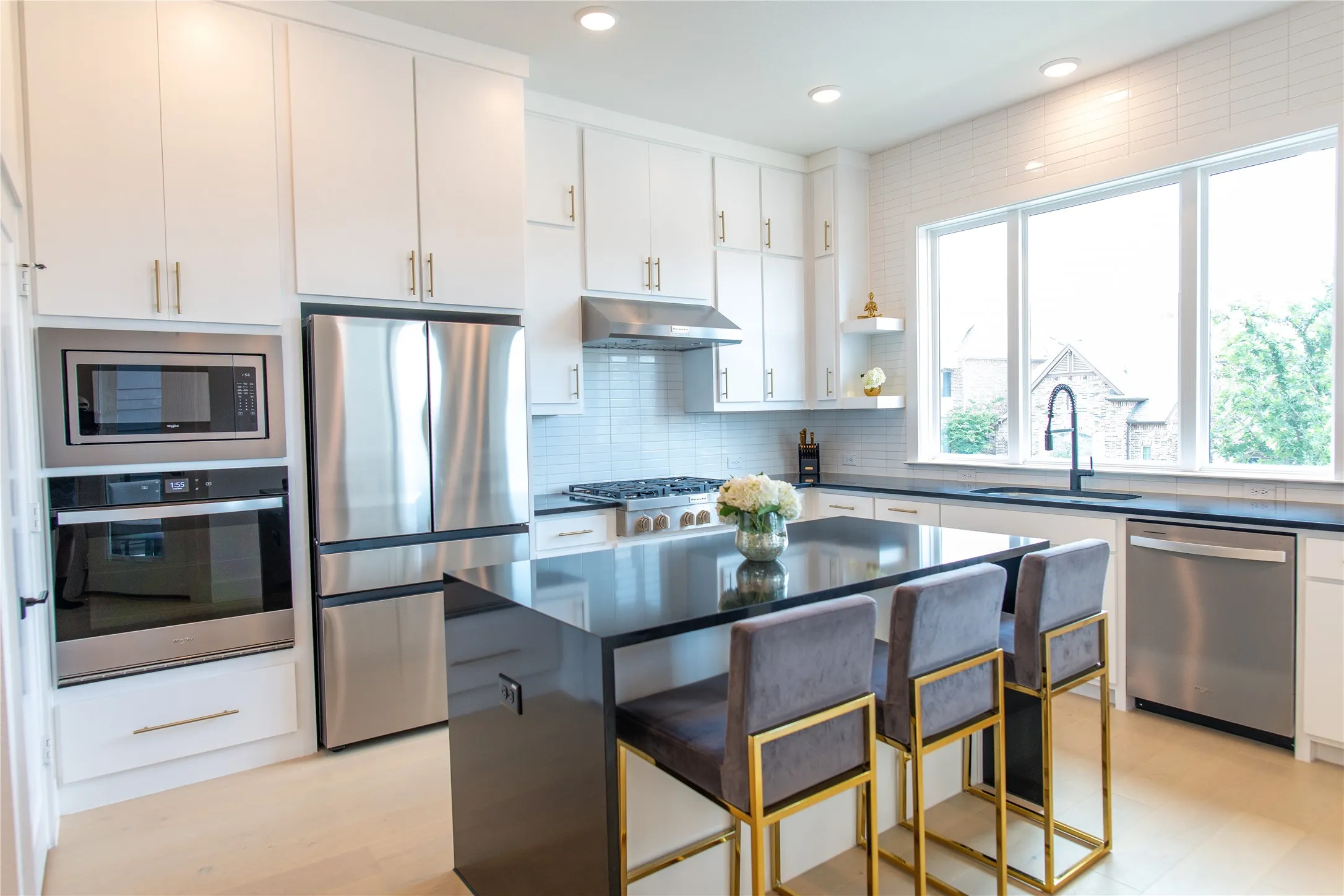 Modern kitchen featuring a black island with counter seating, stainless steel appliances, white cabinetry with gold-tone hardware, a white subway tile backsplash, and light wood-finish flooring