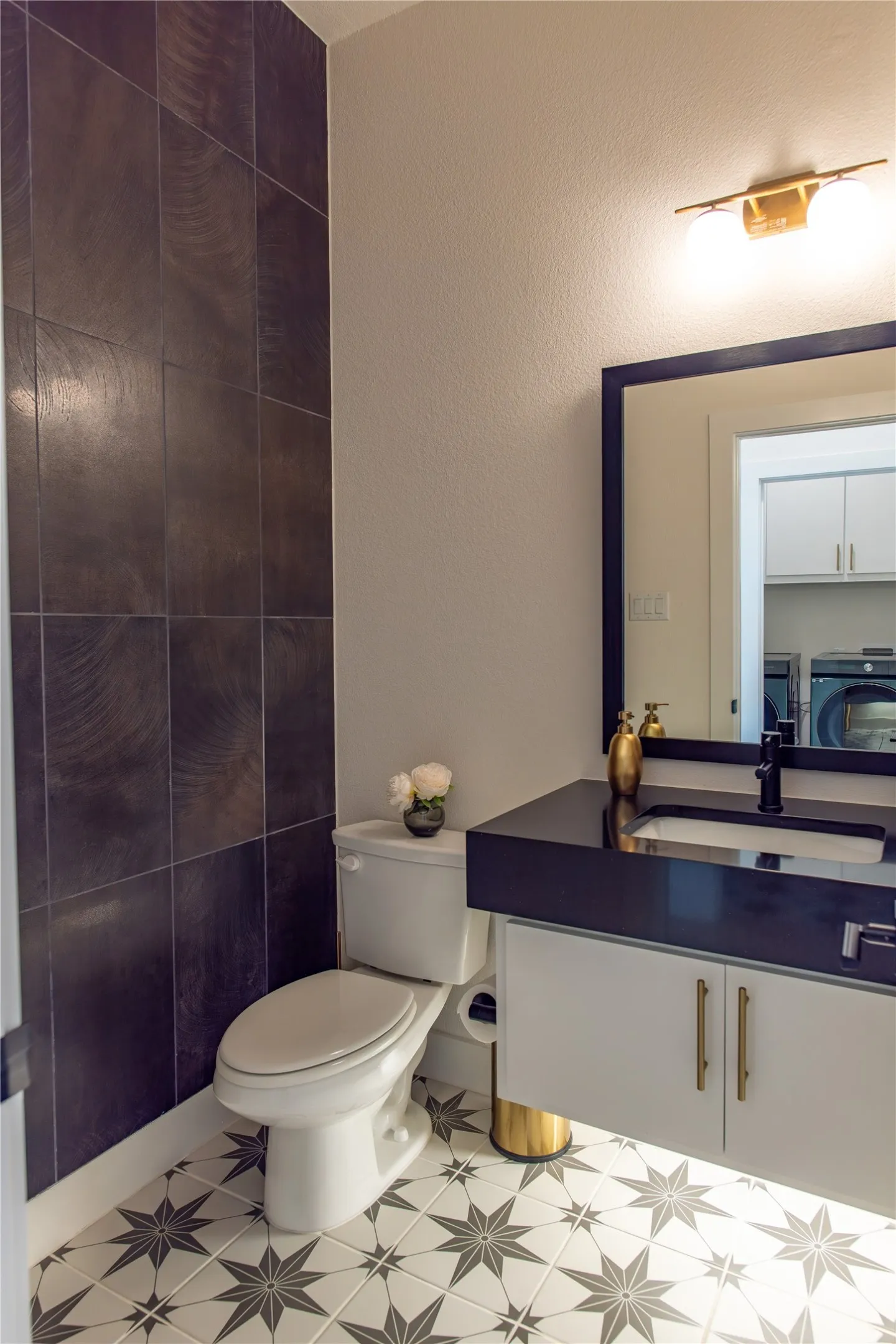 Powder room featuring a patterned floor tile, dark wall tile accent, a white vanity with a black countertop, a black sink faucet, and a gold-toned light fixture