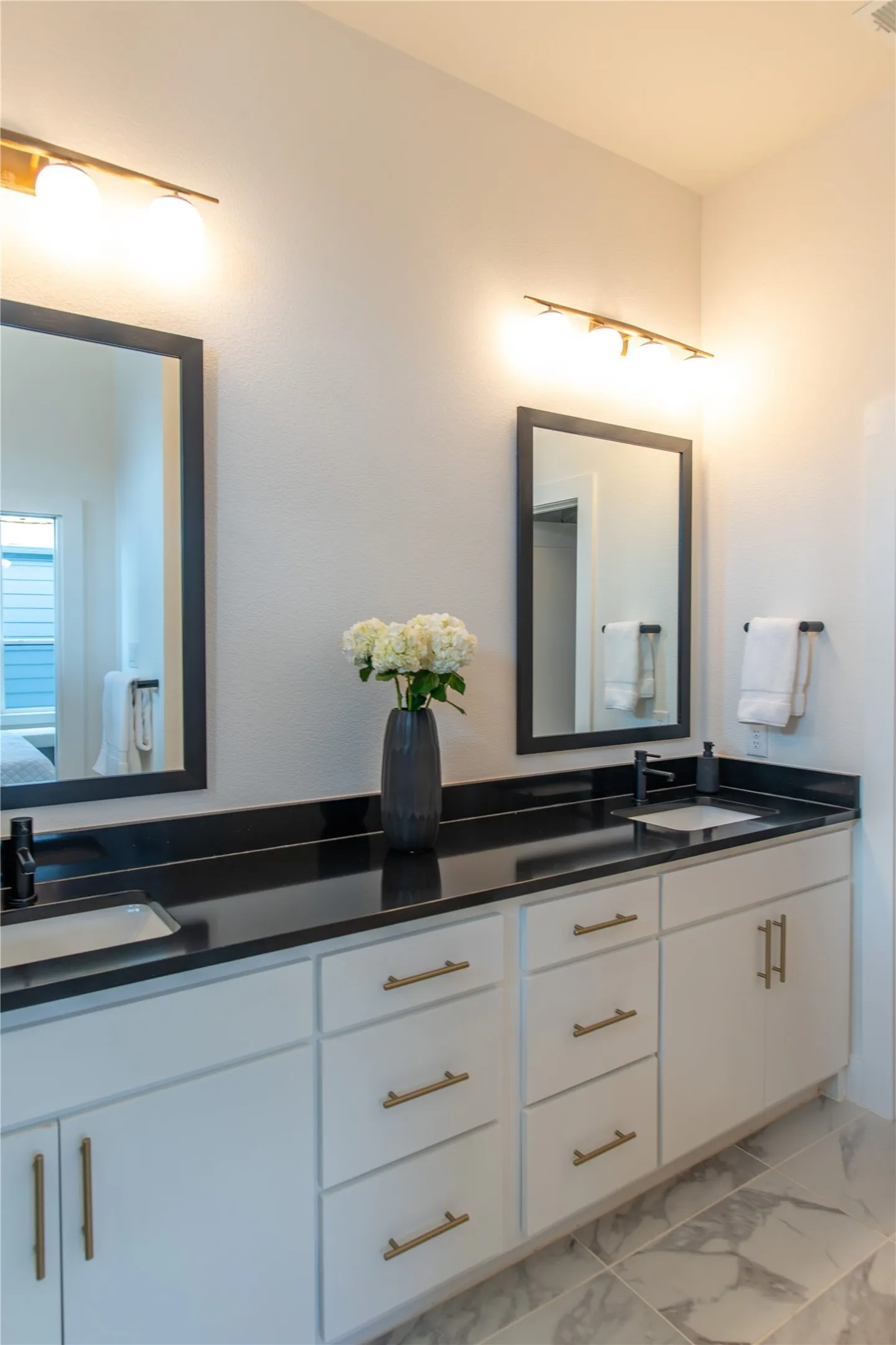 Double vanity featuring dark countertops, white cabinetry with brass hardware, undermount sinks, and wall-mounted light fixtures