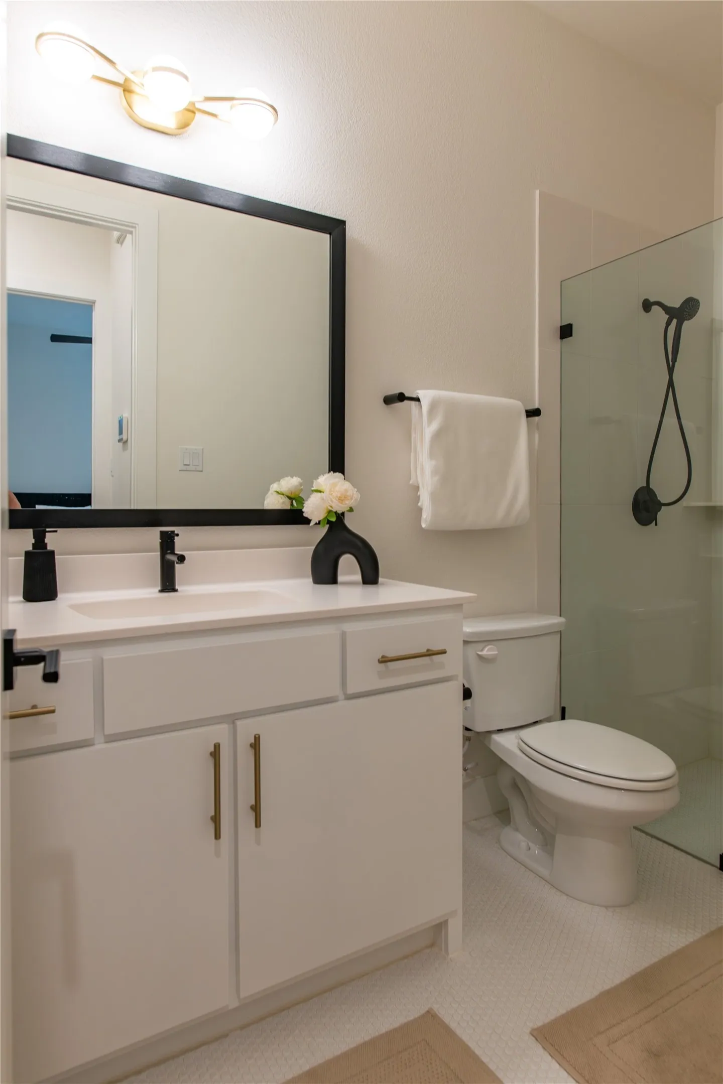Bathroom featuring a white vanity with gold-tone hardware, a rectangular framed mirror, a modern brass light fixture, a toilet, and a glass-enclosed shower with matte black fixtures