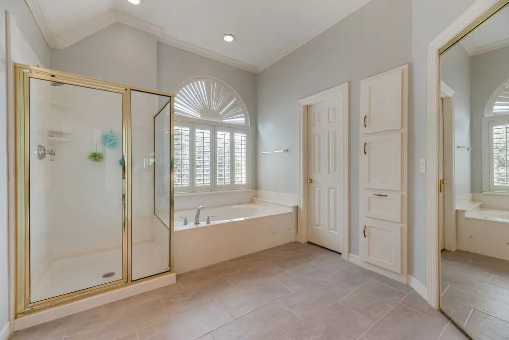 Spacious bathroom featuring a built-in soaking tub with deck-mounted fixtures, an arched window with plantation shutters, and a separate shower with a gold-finish frame