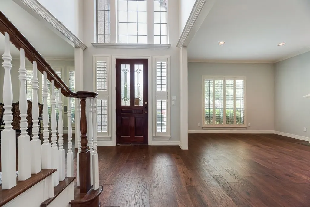 Two-story foyer featuring a dark wood entry door with decorative glass, wood-finish flooring, white plantation shutters, and a wood banister staircase