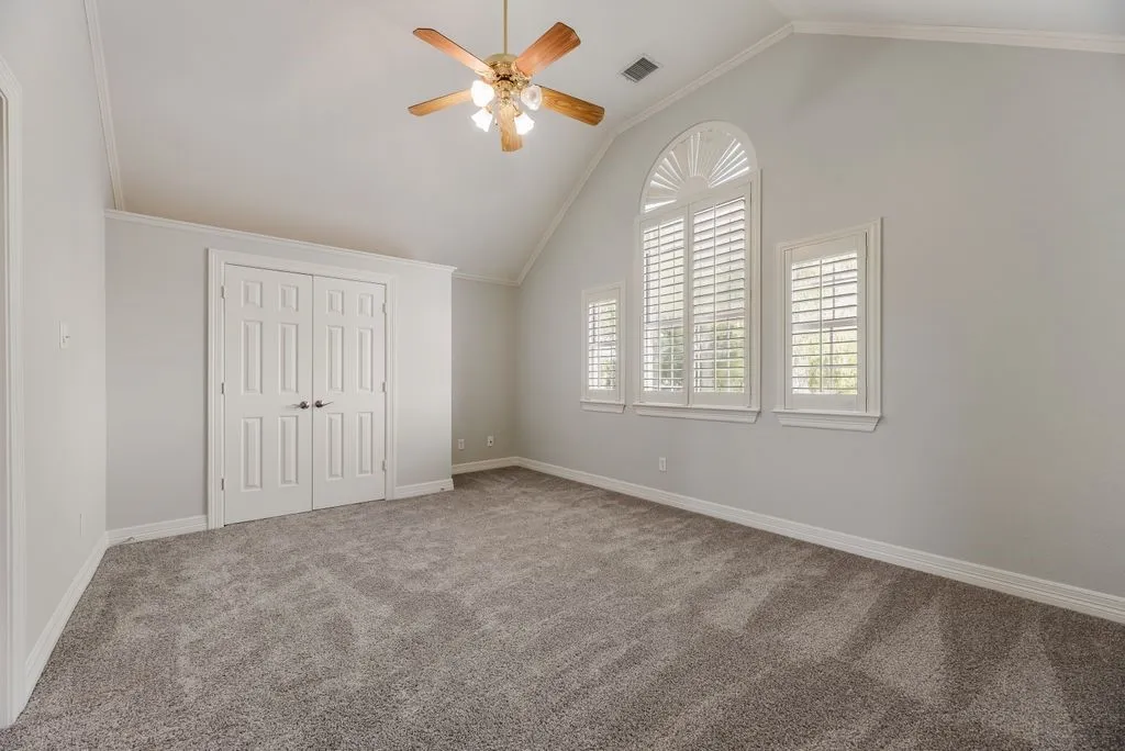 Vaulted ceiling with an overhead fan light fixture, gray carpeting, dual closet doors, and multiple windows featuring plantation shutters