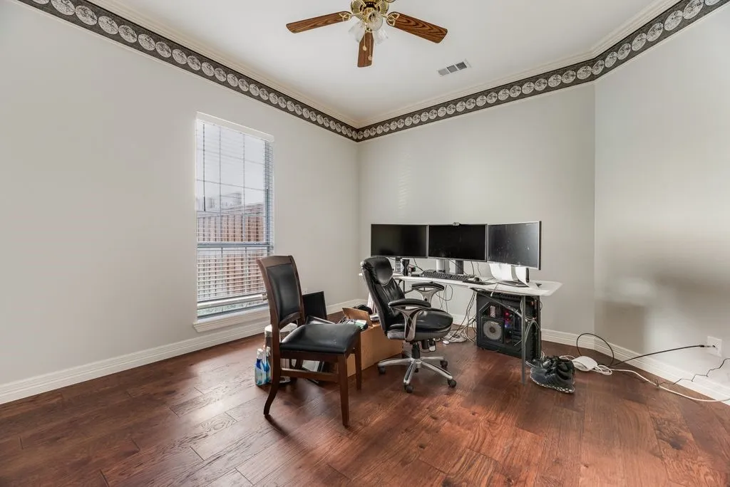 Spacious room featuring rich wood-finish flooring, a ceiling fan fixture, crown molding with decorative trim, a large window with blinds, and light-toned walls