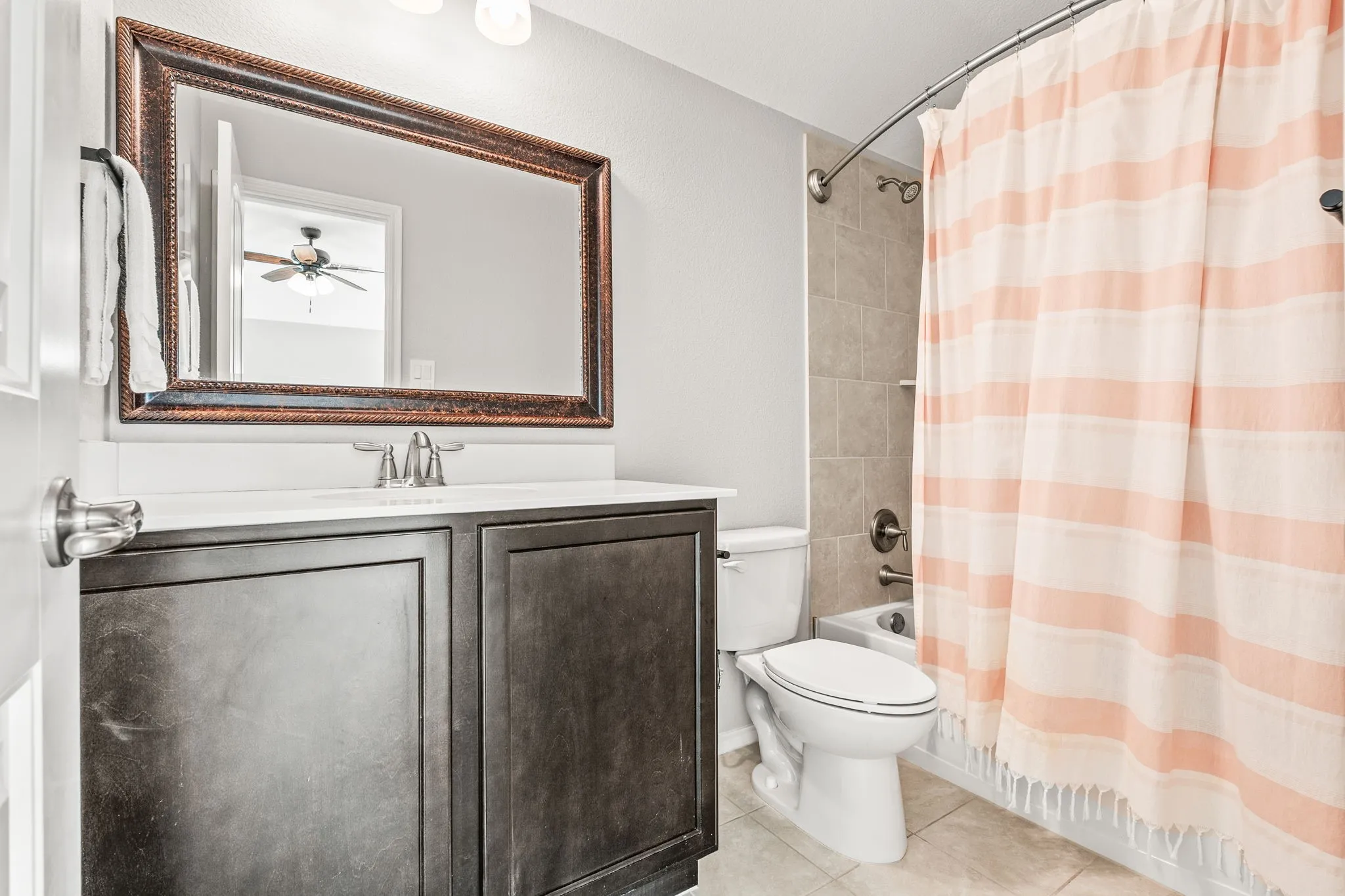 Bathroom featuring a dark wood-finish vanity with a white countertop, a bronze-framed mirror, a toilet, a bathtub with tiled surround, and floor tiling