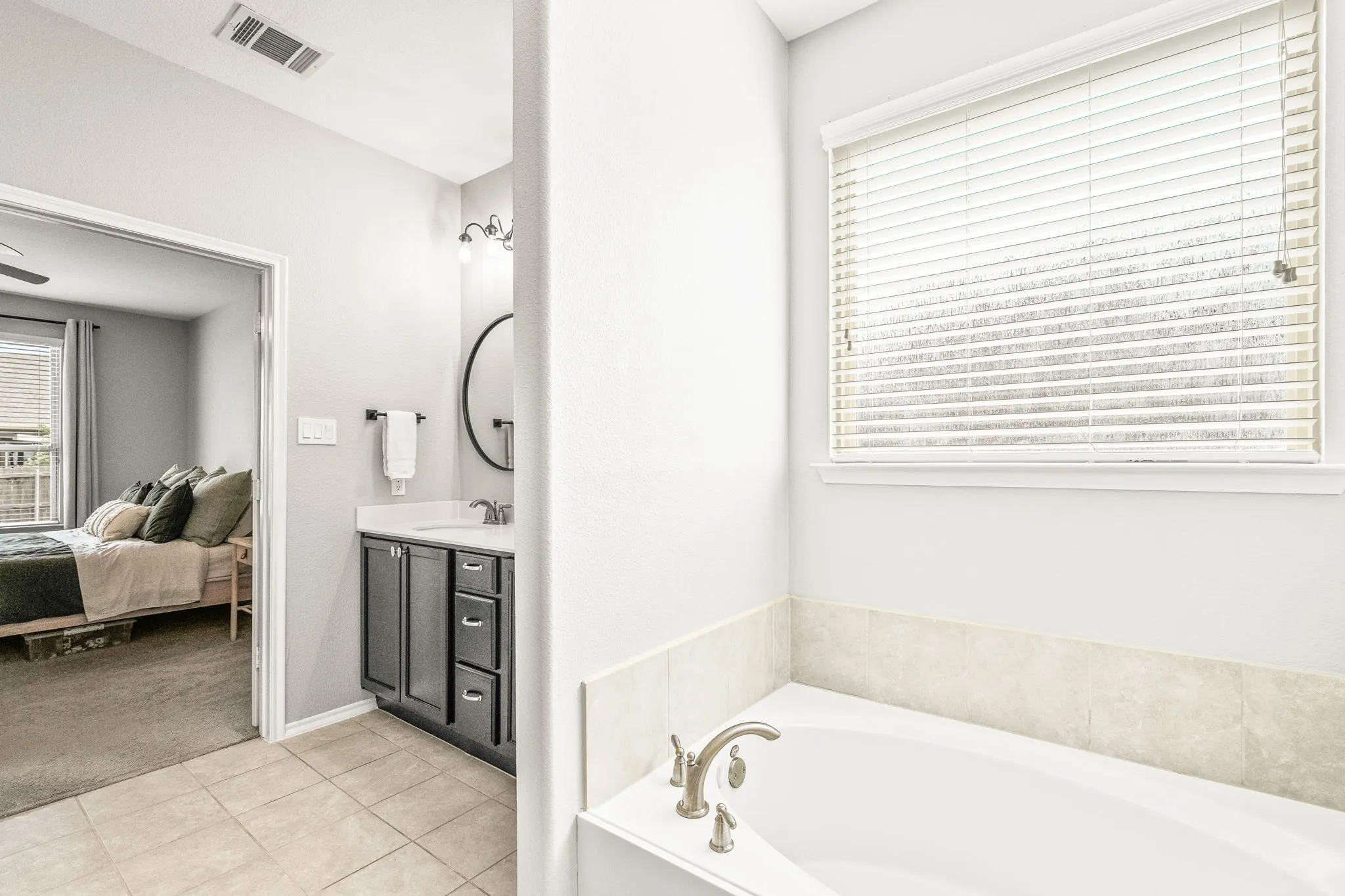Soaking tub with tile surround, paired with a single vanity featuring a dark wood-finish cabinet and a circular mirror