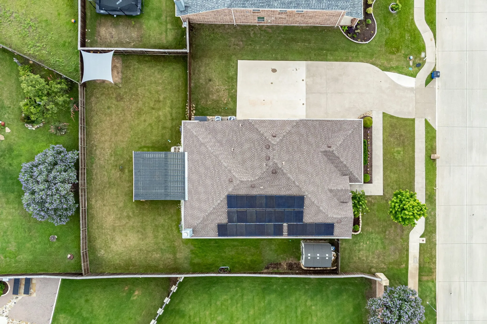 Overhead property perspective showcasing a substantial residential roofline with integrated solar panels, a concrete driveway, a paved walkway, a manicured lawn, and a fenced backyard