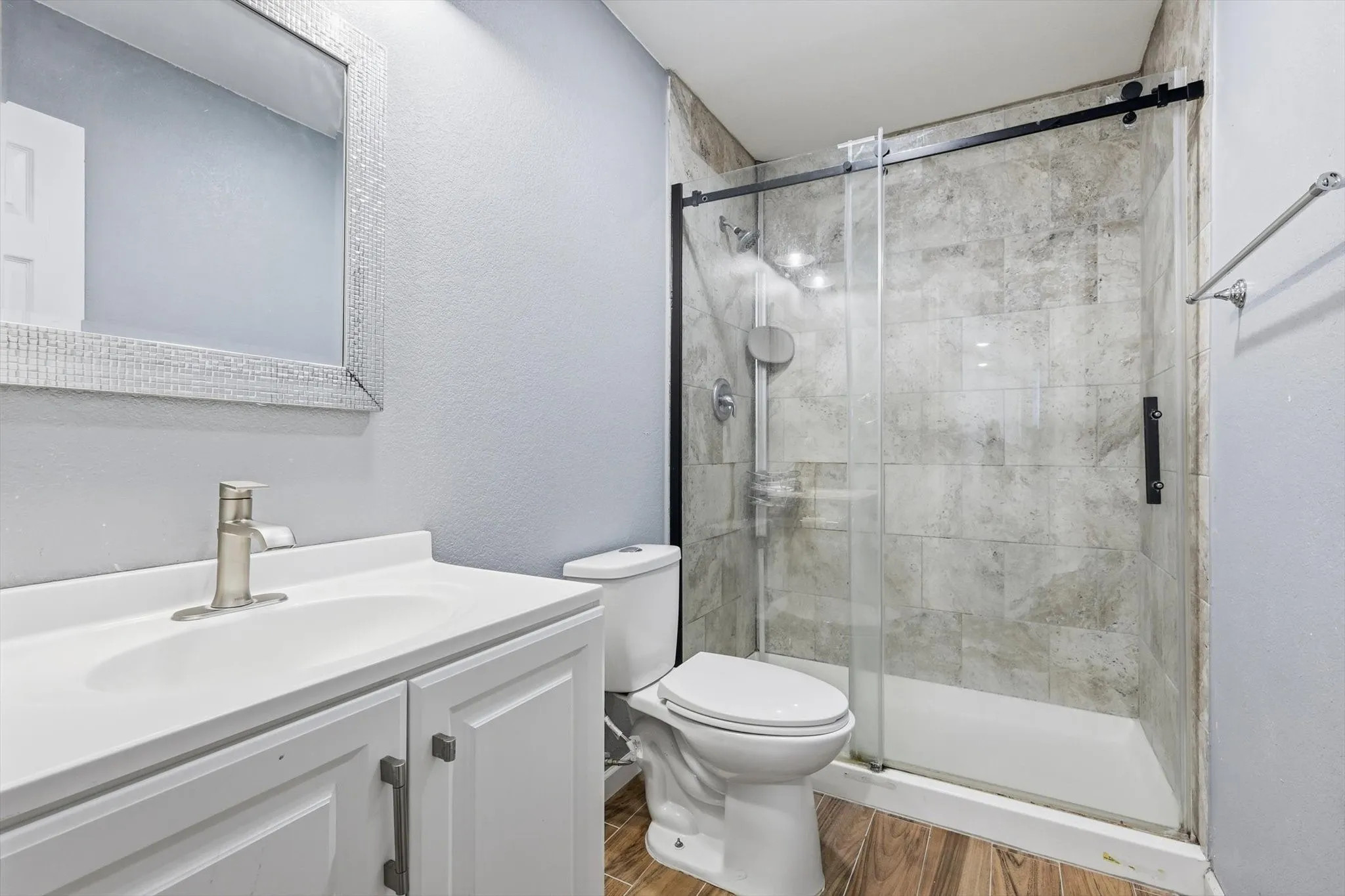Bathroom featuring a glass-enclosed shower with tile surround, a white vanity with an integrated sink, a framed mirror, and wood-finish flooring