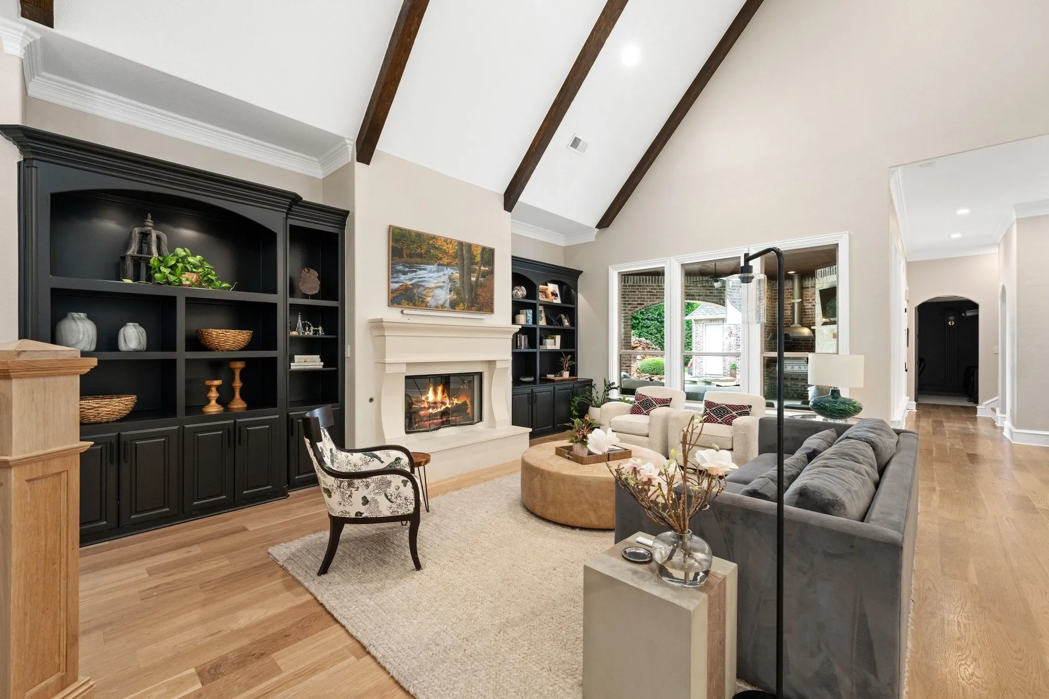 Vaulted ceiling with exposed wood beams, a stone-surround fireplace with mantel, built-in cabinetry, large windows, and light wood-finish flooring