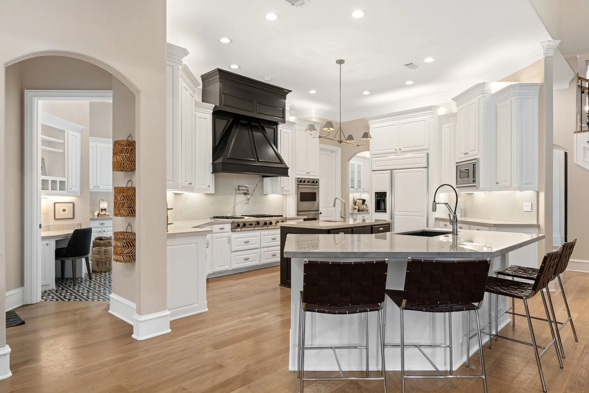 Spacious kitchen featuring light wood-finish flooring, white cabinetry, a large central island with seating, a dark wood-finish range hood, and a desk area with patterned tile flooring