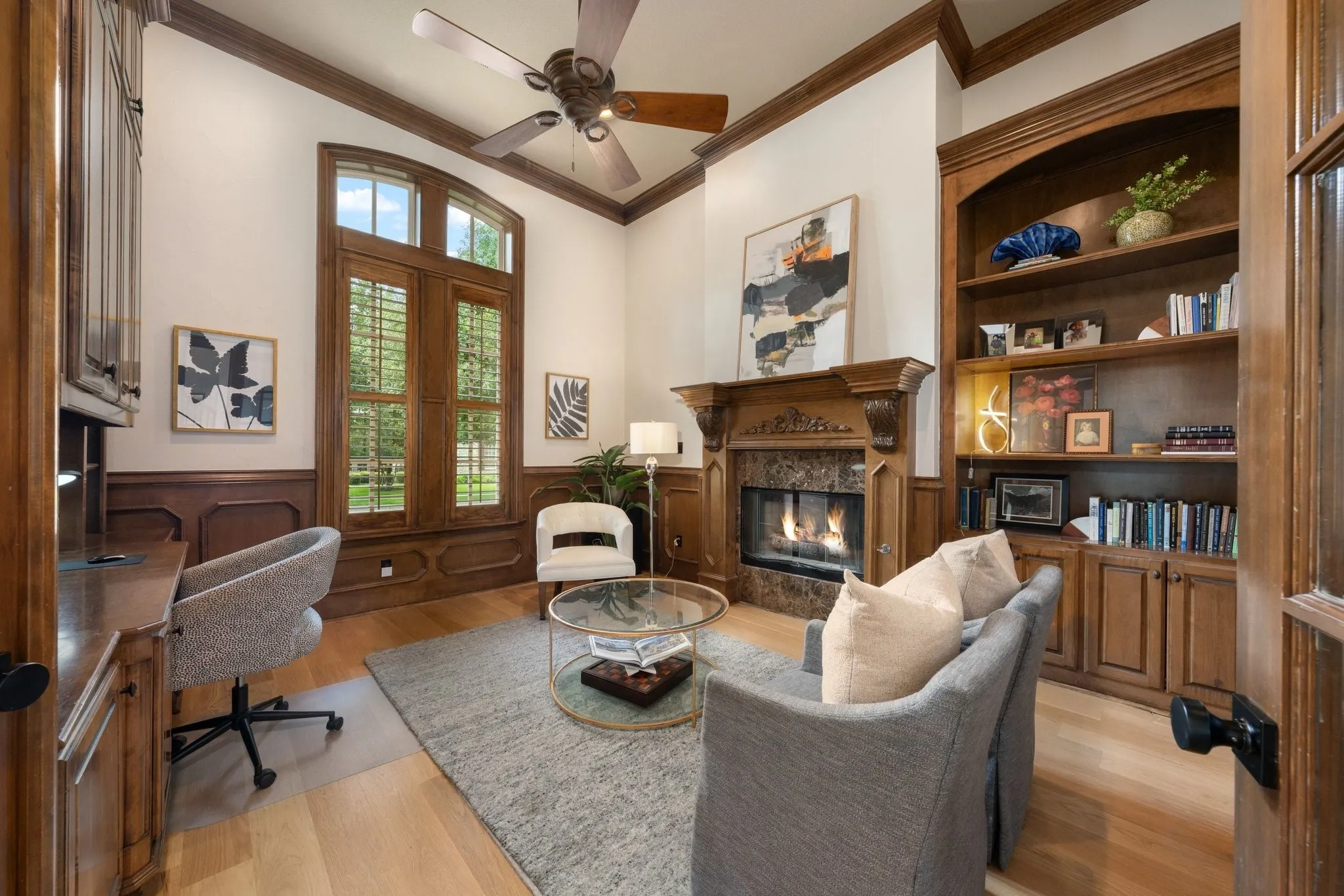 Hardwood flooring throughout this room featuring a fireplace with a stone surround and ornate wood mantel, built-in shelving with cabinetry, a built-in desk, and a large window with wood shutters