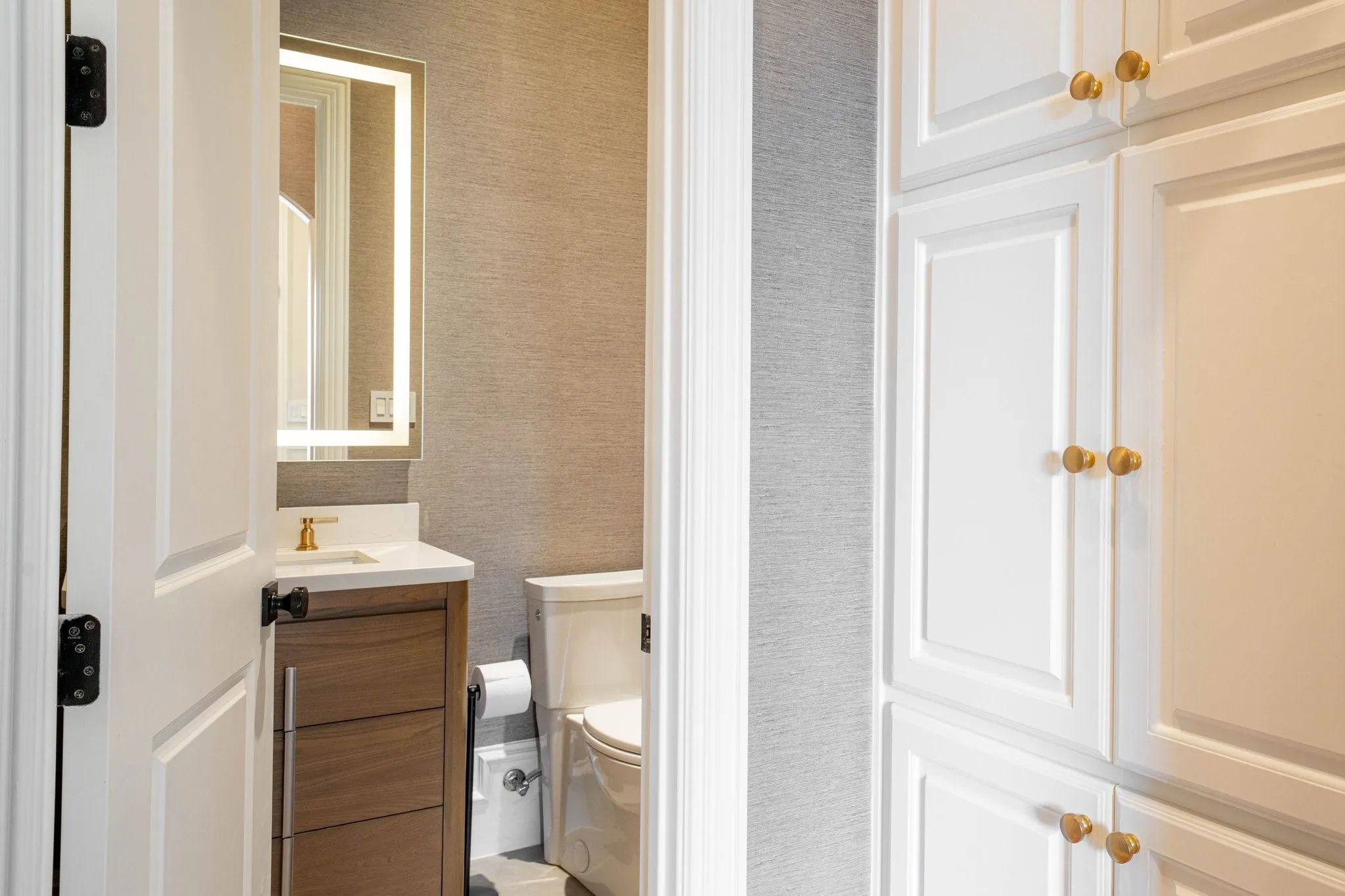 Powder room featuring a wood-finish vanity with a white countertop and a gold-finish faucet, an illuminated mirror, and textured wall covering