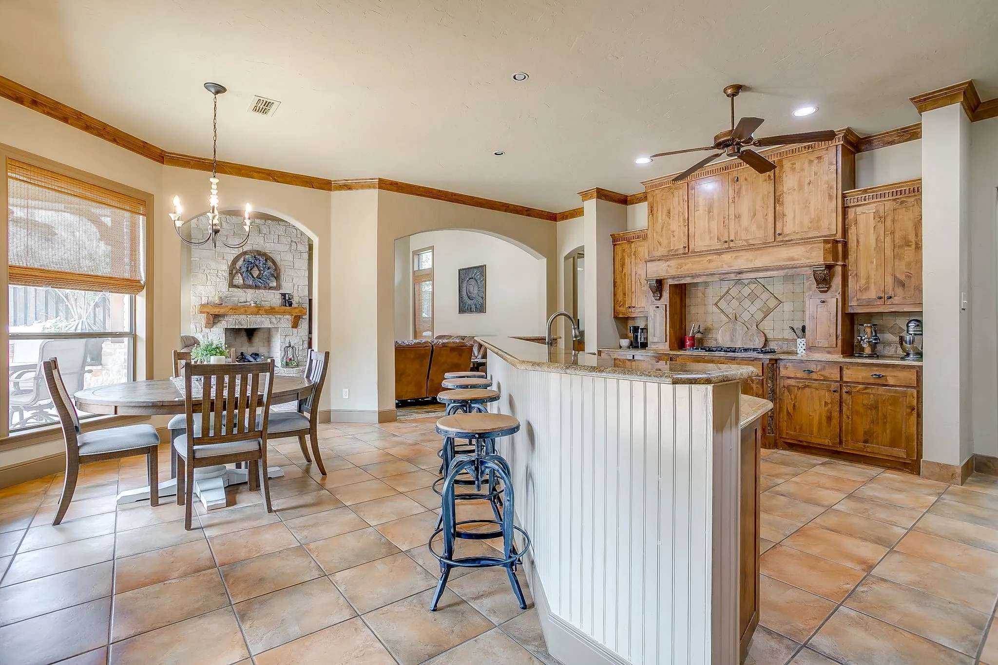 Expansive kitchen and dining area featuring ceramic tile flooring, wood cabinetry, a central island with bar seating, an arched stone fireplace, and a large window with natural light