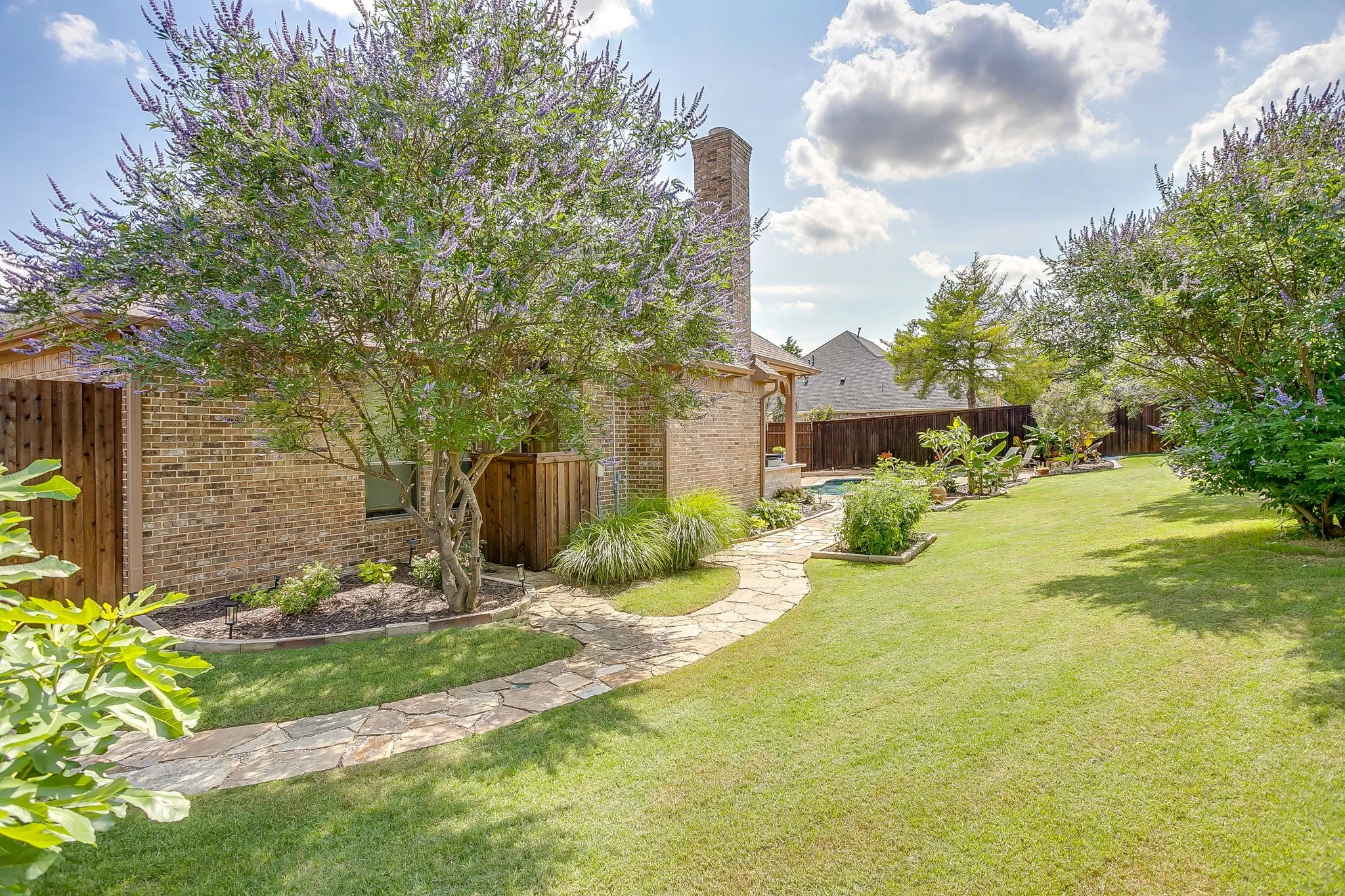 Expansive backyard featuring a manicured lawn, flagstone pathway, mature landscaping, brick exterior, and wood fencing