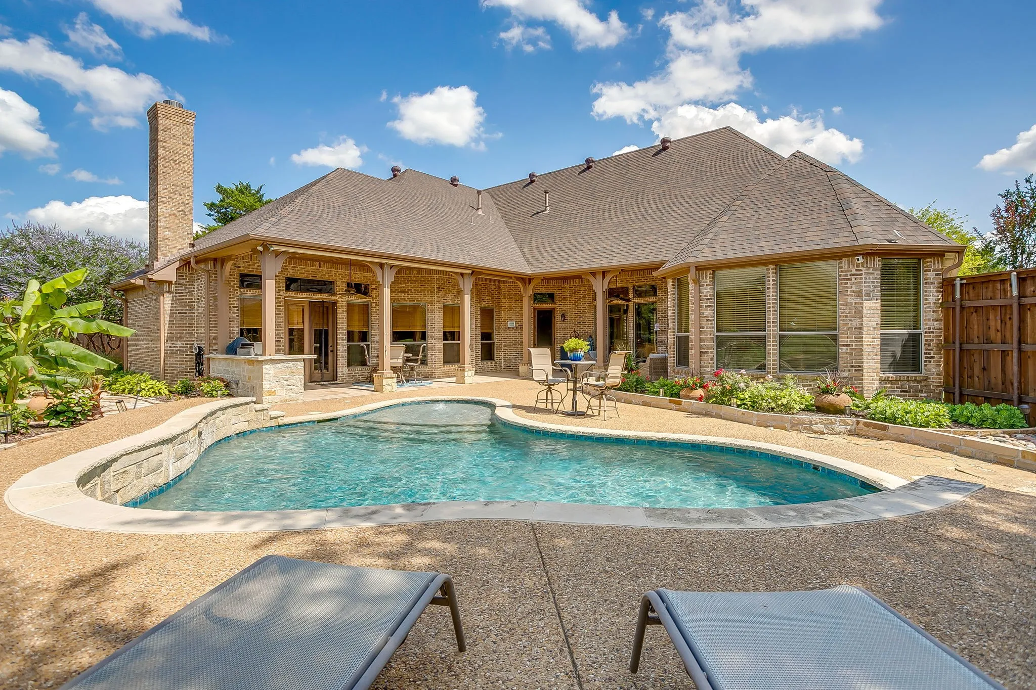 Outdoor oasis featuring a freeform swimming pool with integrated spa, expansive exposed aggregate decking, and a covered patio with columns