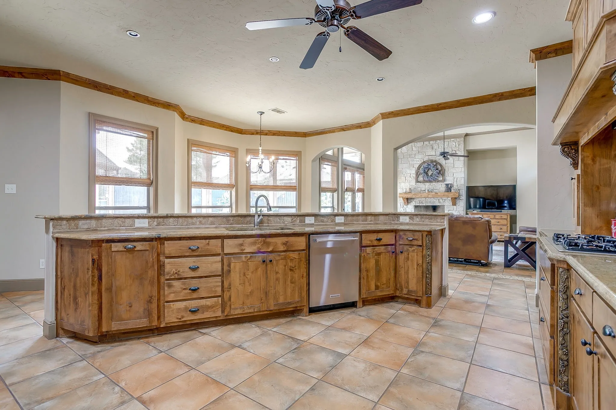 Spacious kitchen featuring wood cabinetry, stone countertops, a large island with sink, a stainless steel dishwasher, and tile flooring