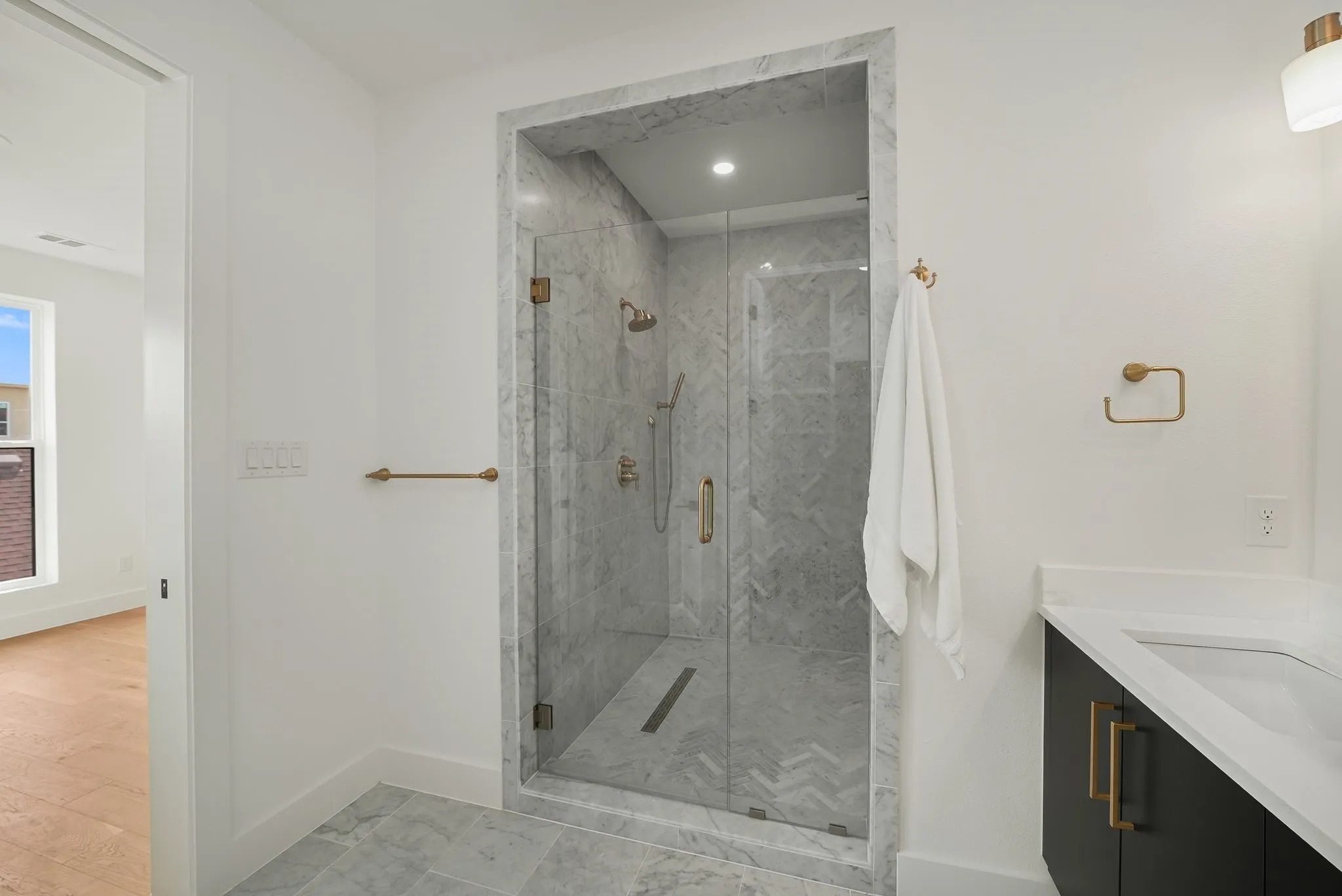 Glass-enclosed shower with a rainfall showerhead, marble-style wall tiles in a herringbone pattern, coordinating marble-style floor tiles, and a linear drain