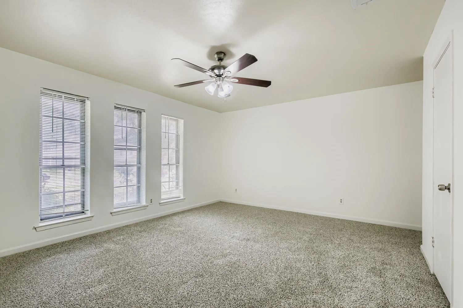 Spacious room featuring three vertical windows, a ceiling fan with light fixture, light neutral walls, and textured carpet flooring