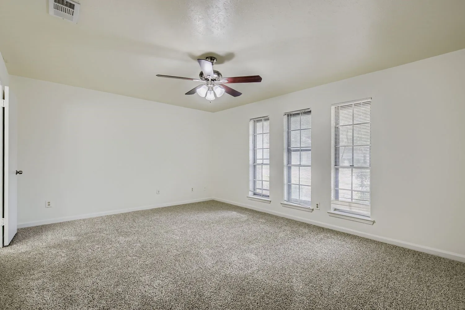 Spacious interior room featuring light-toned walls, neutral-toned carpet flooring, three vertical windows with blinds, and a ceiling fan with light fixture