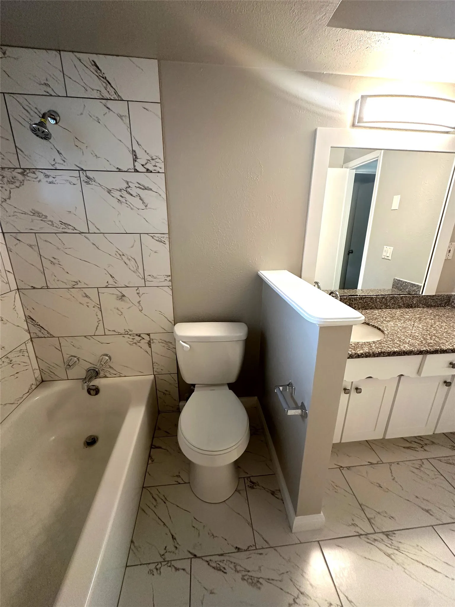Bathroom featuring a white porcelain tub with a shower fixture, white and grey marble-look wall tile, a white toilet, and marble-look floor tile