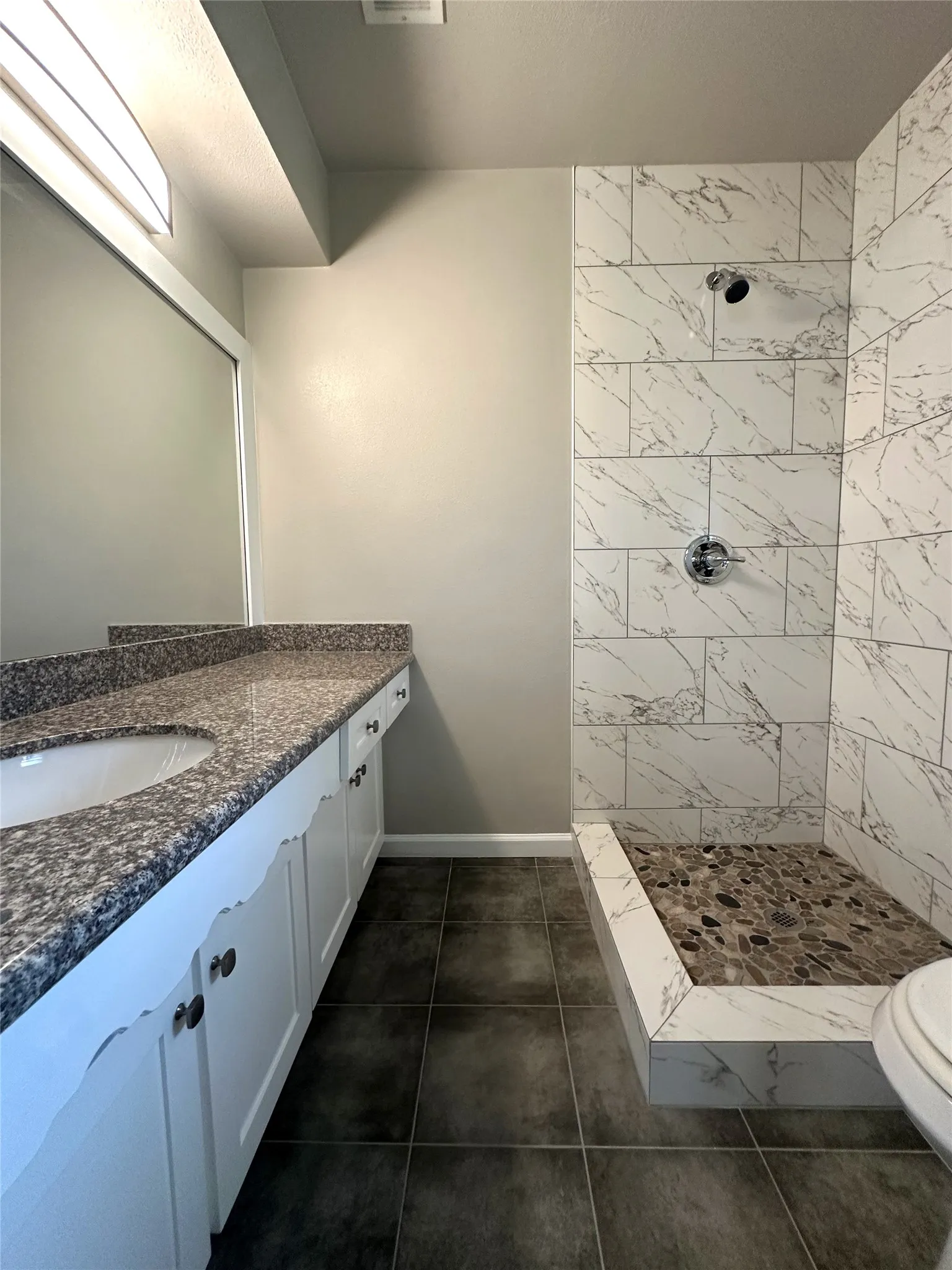 Bathroom featuring a dual vanity with a speckled countertop, white cabinetry with dark hardware, a walk-in shower with patterned wall tile, and a pebble-stone shower floor
