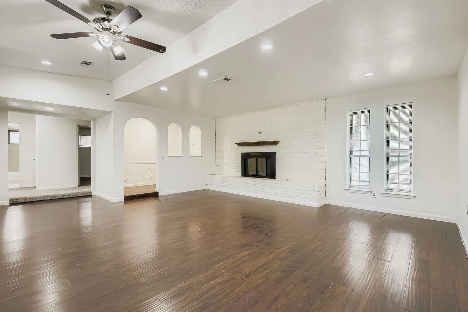 Expansive living area featuring rich wood-finish flooring, a painted brick fireplace with a wood mantel, and arched wall niches