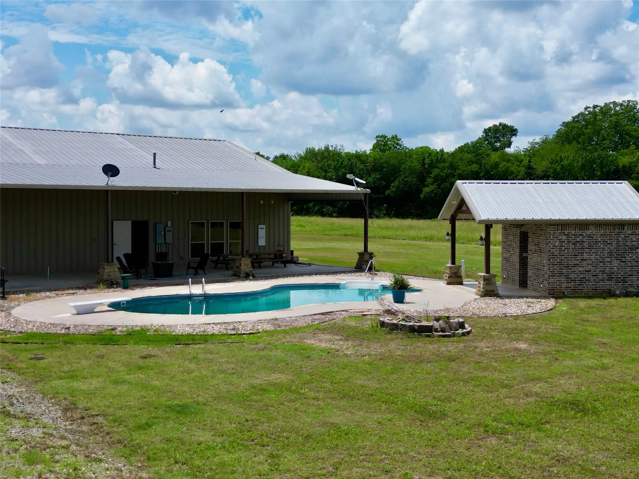  Covered patio Expansive backyard oasis featuring a freeform swimming pool with a diving board, surrounding concrete patio, and a stone-clad pool house with a metal roof