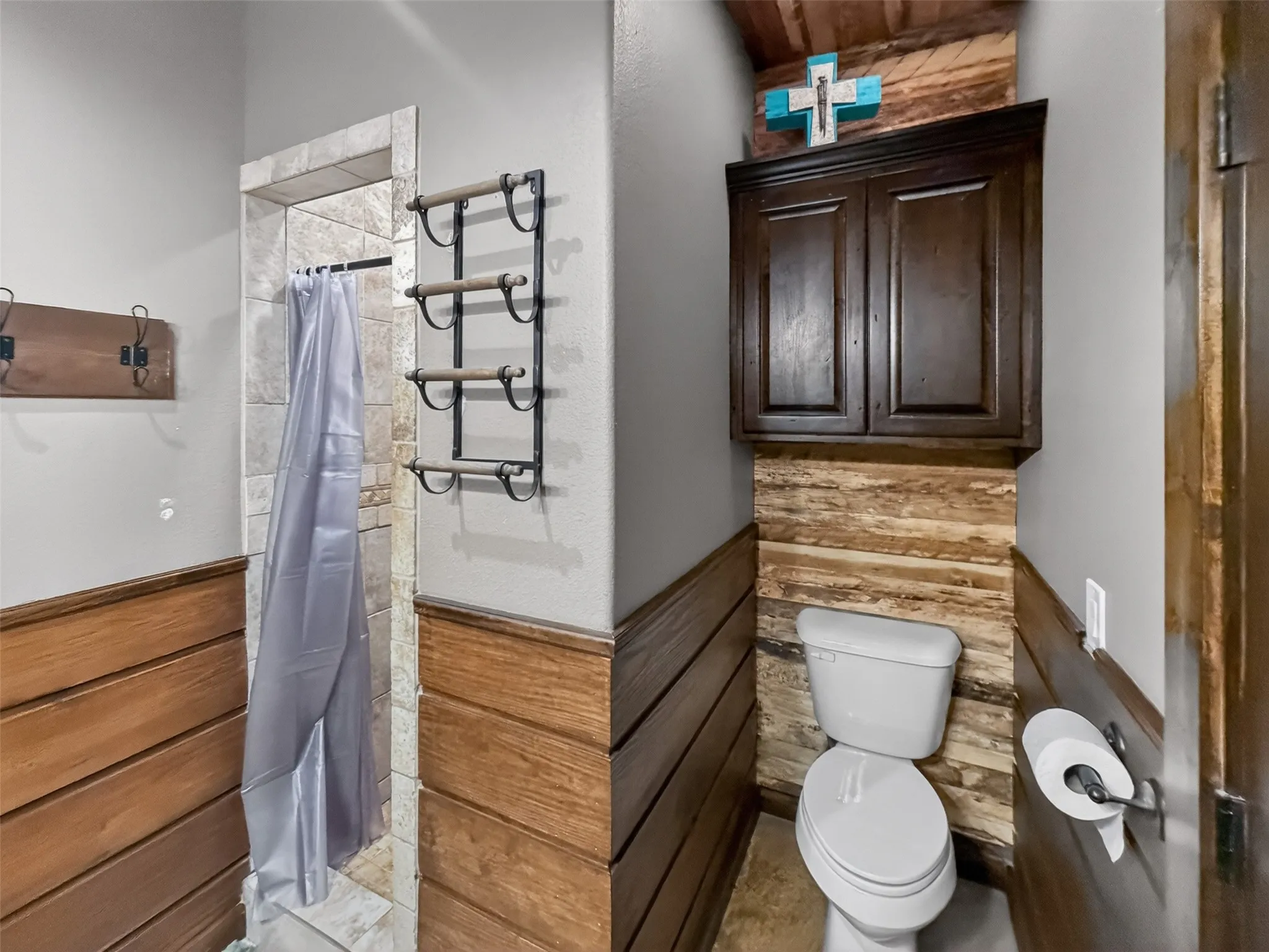 1st floor bathroom featuring a built-in shower with tile surround, wood-finish wainscoting, and a dark wood-finish wall cabinet above the commode