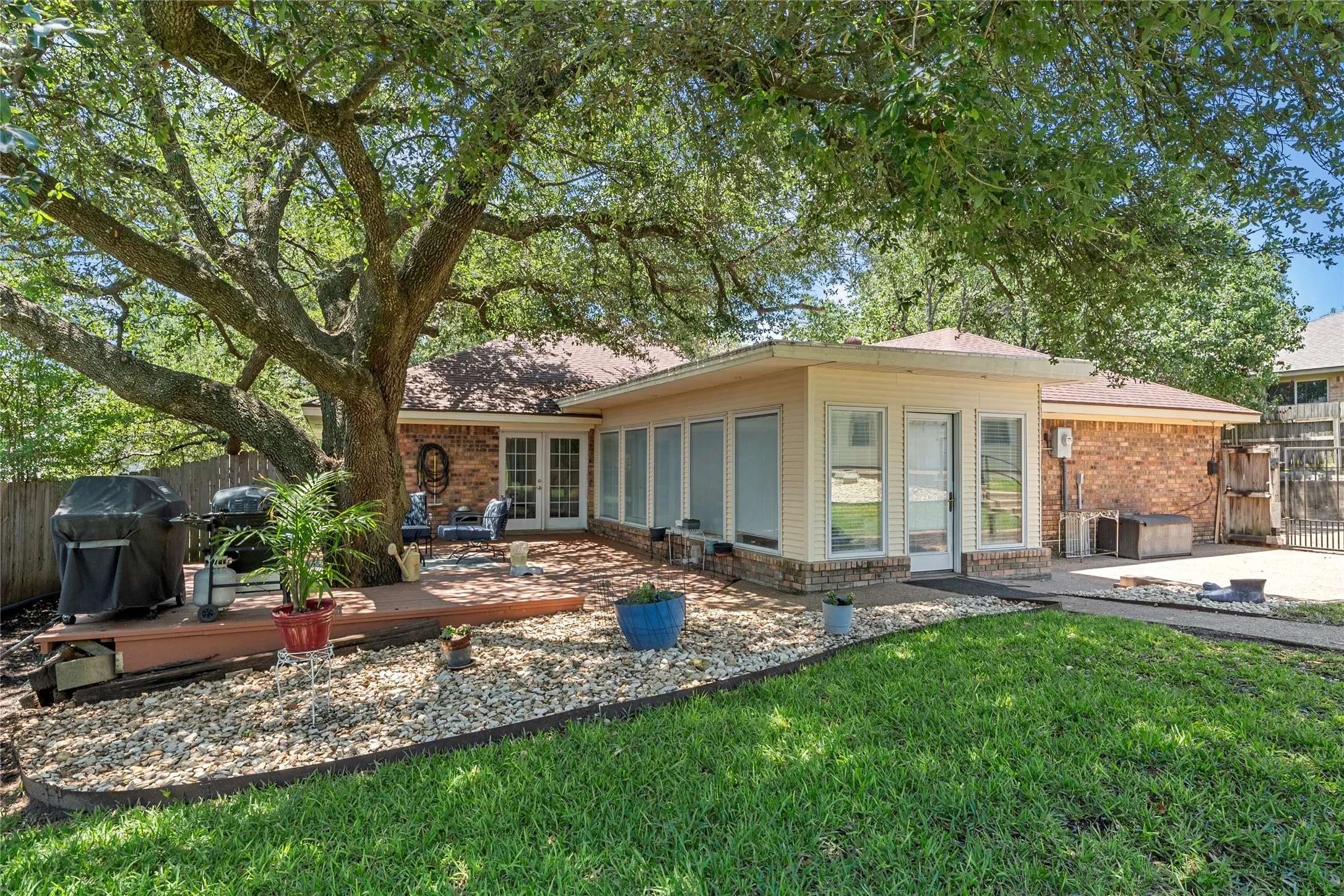 Spacious backyard featuring a substantial mature tree, a generous wood-finish deck, a rock garden, a manicured lawn, and a sunroom addition with multiple windows