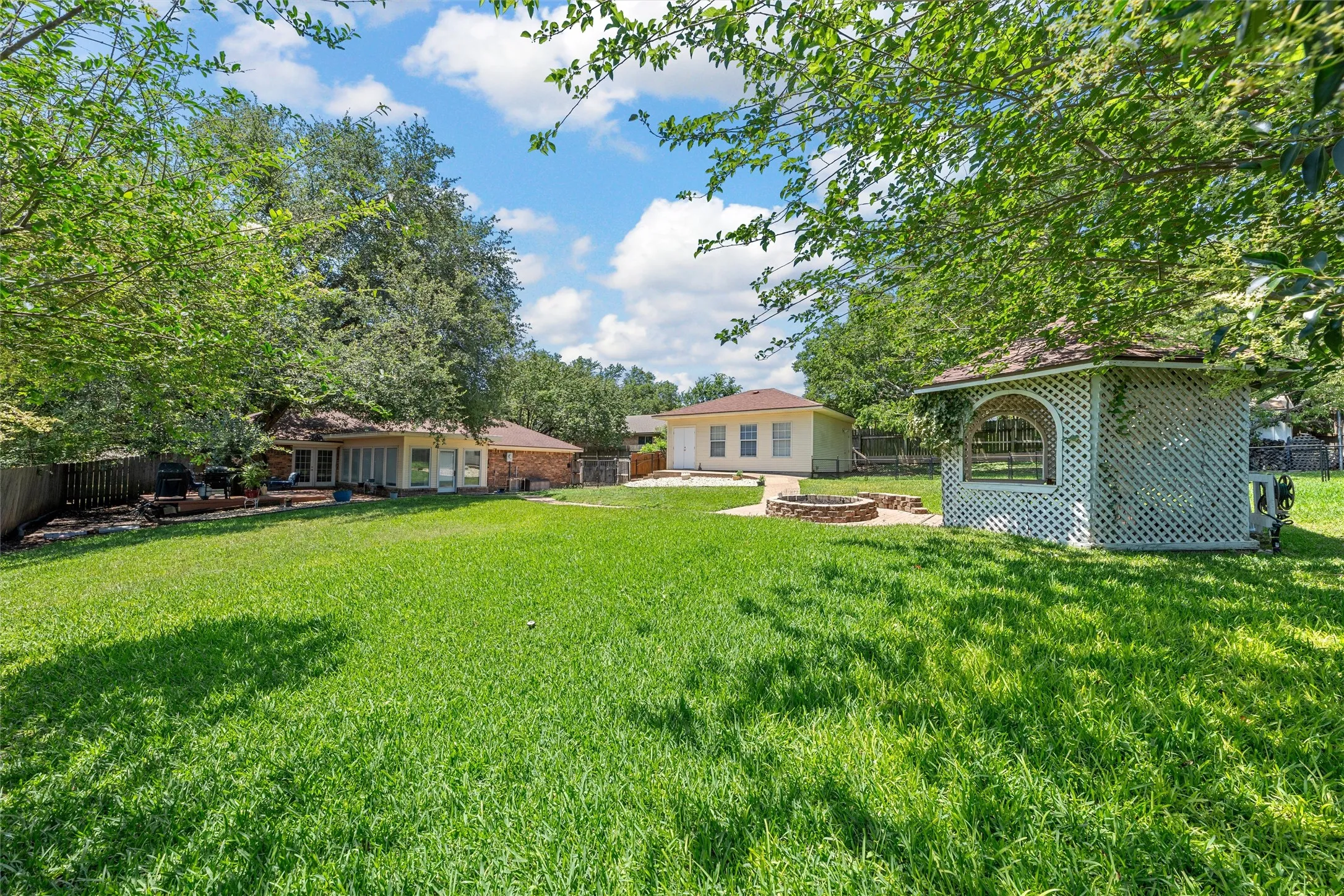Expansive grassy yard featuring a white lattice gazebo, stone fire pit, and a rear building with multiple windows