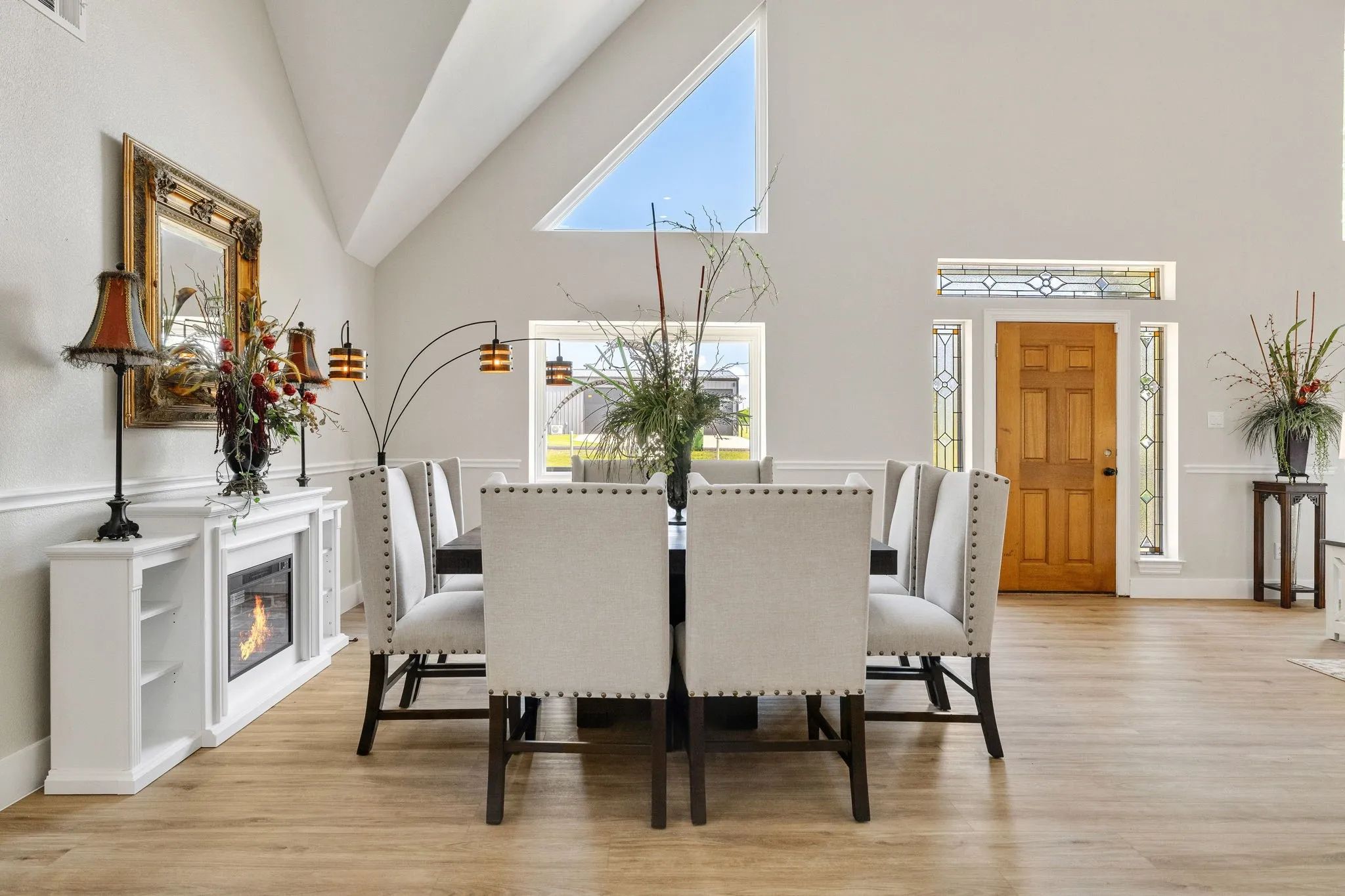 Open-concept dining area featuring vaulted ceilings, wood-finish flooring, a built-in electric fireplace with mantel, and a natural wood entry door with stained glass sidelights