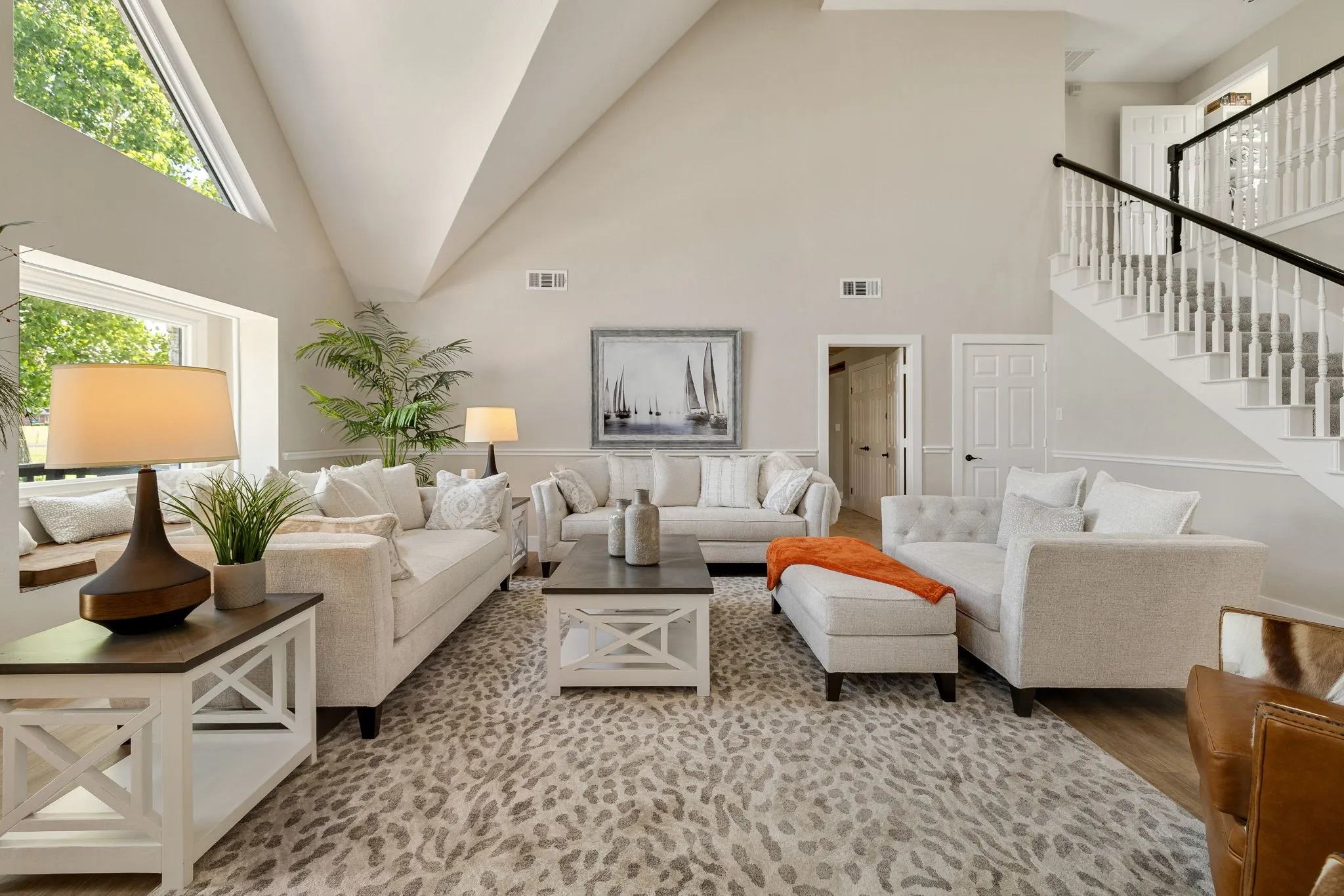 Spacious living area featuring vaulted ceilings, abundant natural light from large windows and an additional transom window, a built-in window bench, a grand staircase with white spindles and a dark handrail, and light wood-finish flooring