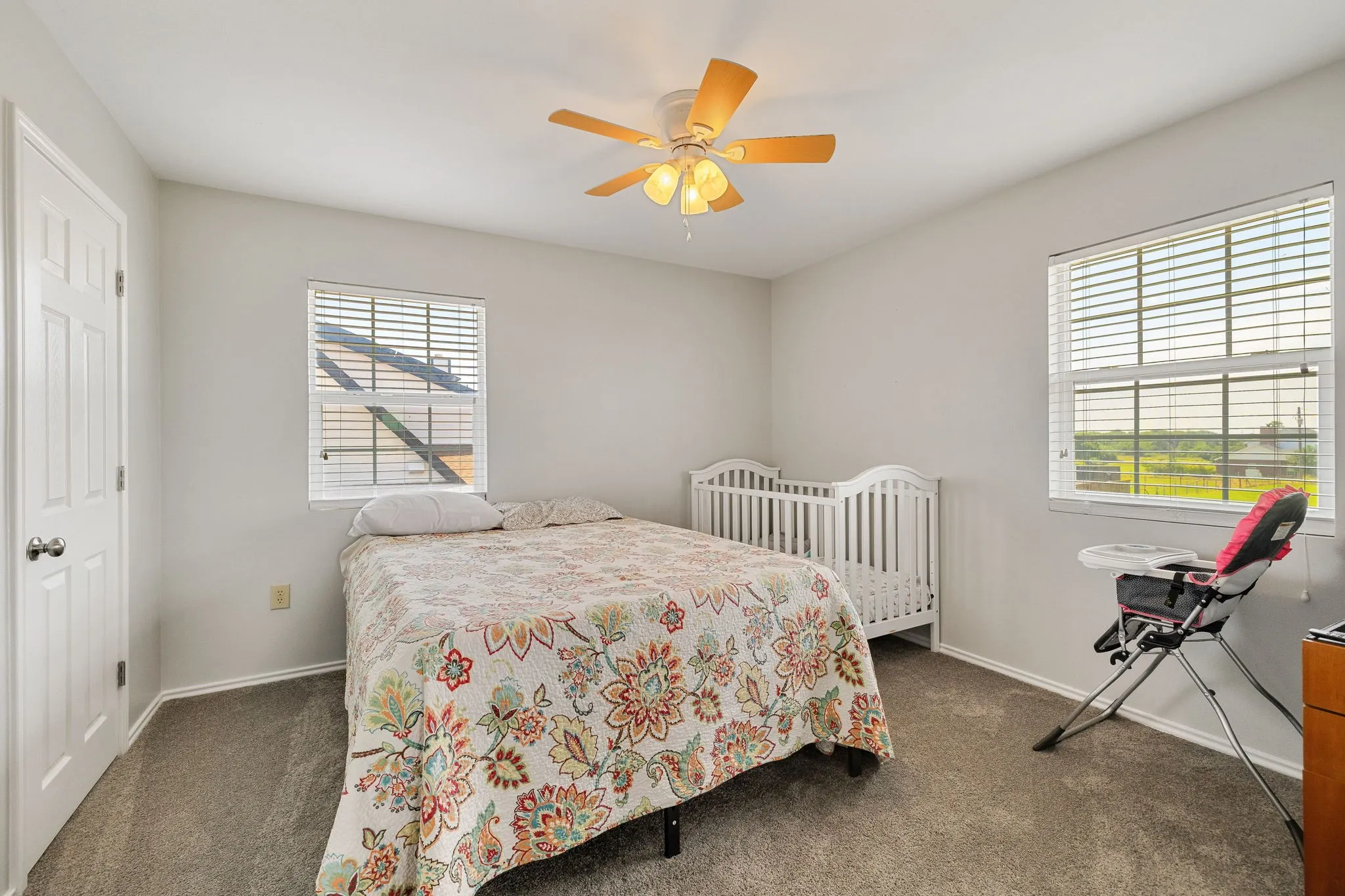 Carpeted interior room featuring light gray walls, two windows with horizontal blinds, a white paneled door, and a ceiling fan with light fixture