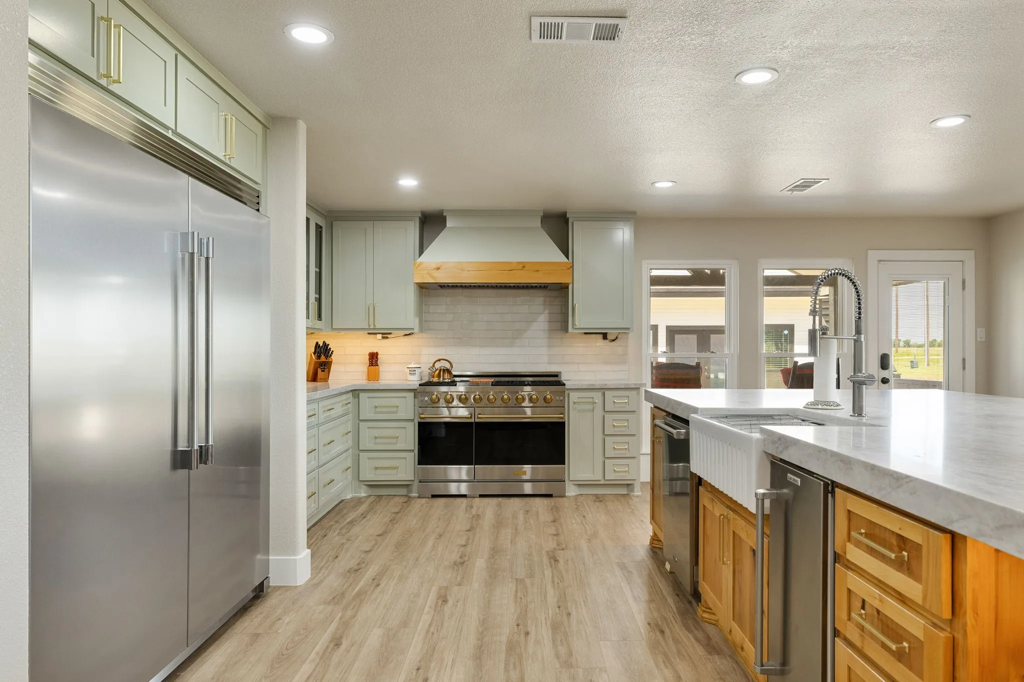 Gourmet kitchen featuring a large stainless steel refrigerator, professional-grade range, custom light green cabinetry, wood-finish flooring, and a spacious island with a farmhouse sink