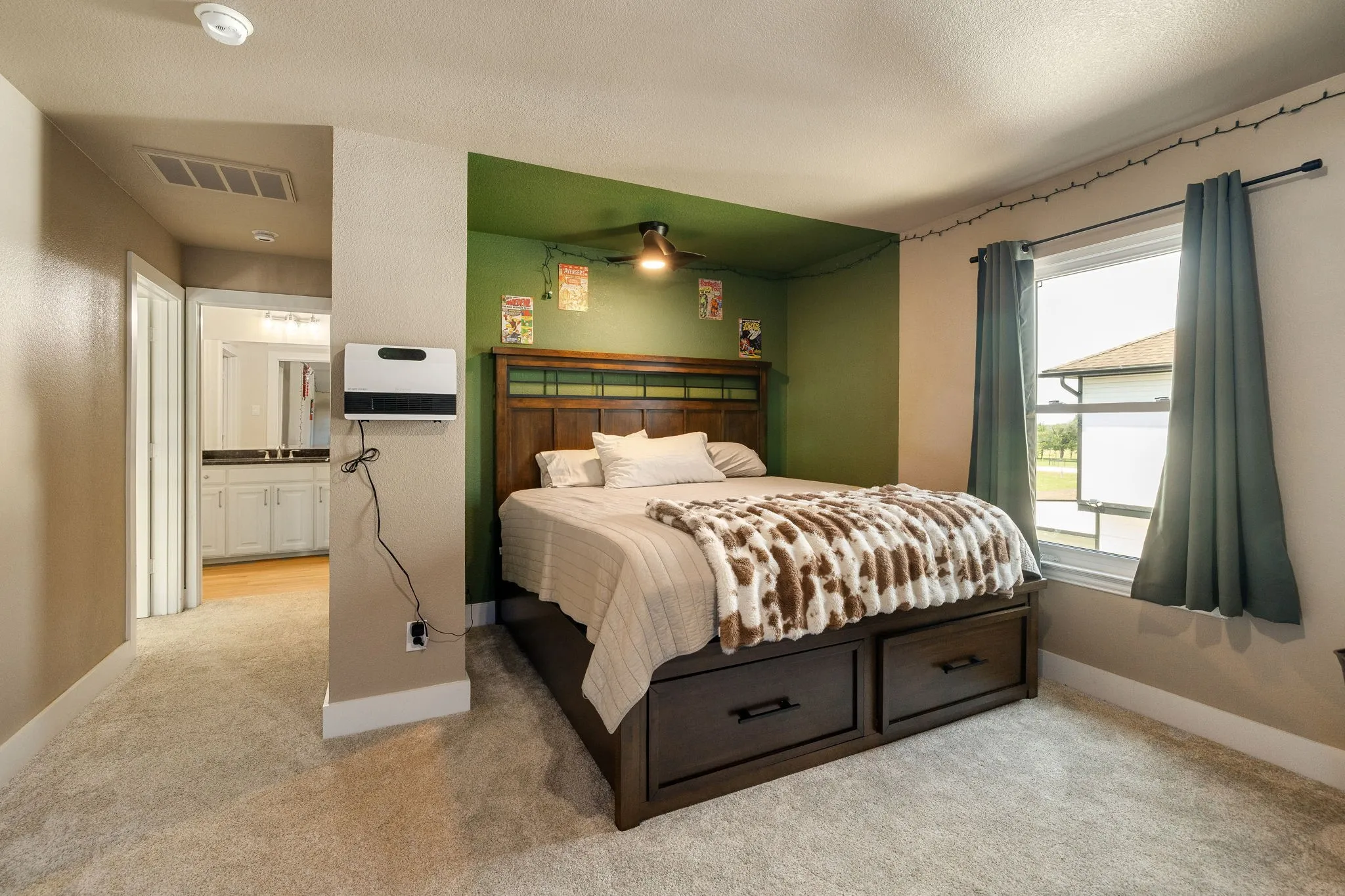 Carpeted bedroom featuring a dedicated bed nook with a ceiling fan, a large window with privacy drapes, and access to a bathroom with a dual vanity
