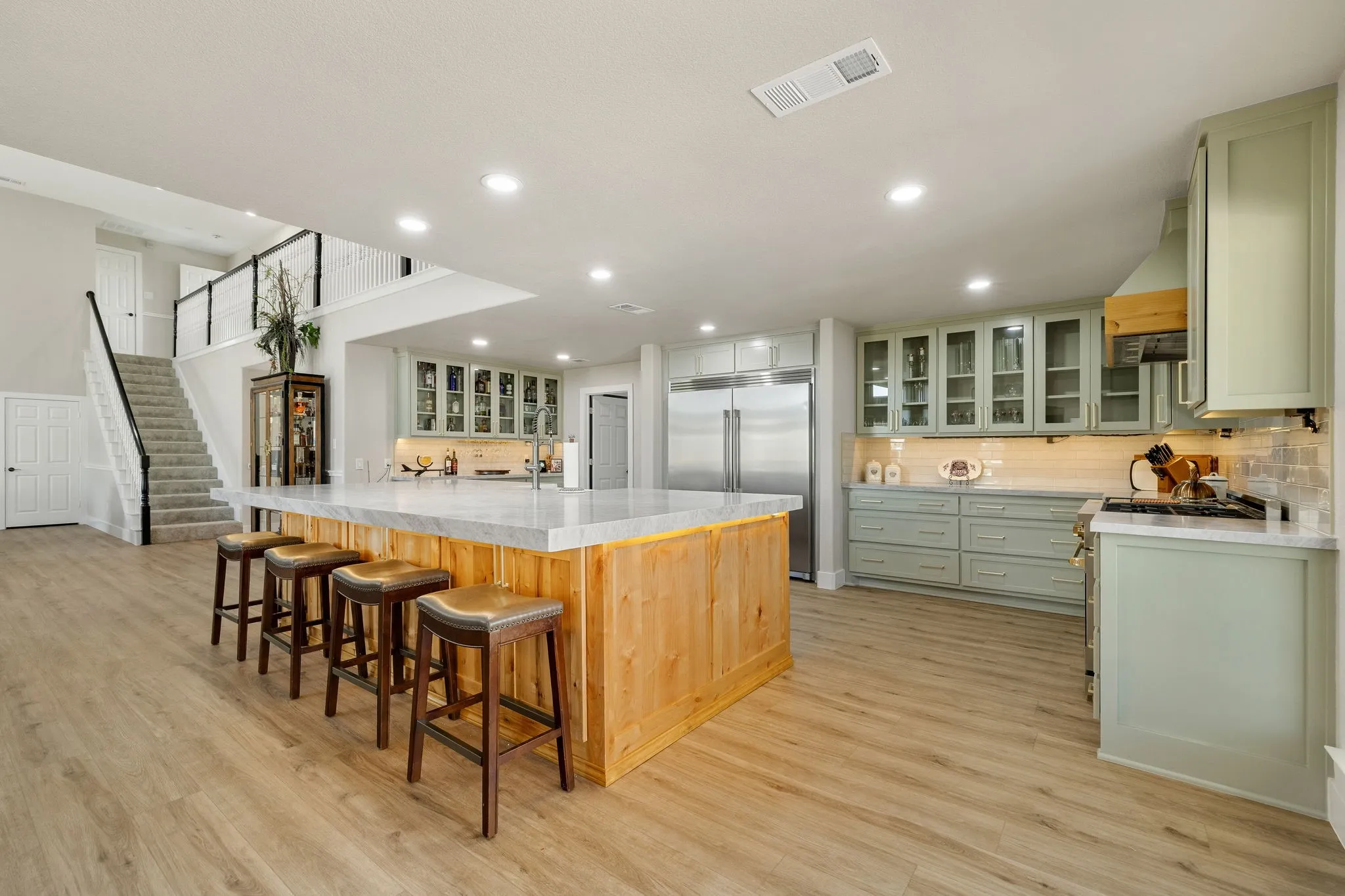 Expansive kitchen featuring a large wood-paneled island with seating, light green cabinetry, stainless steel appliances, and wood-finish flooring