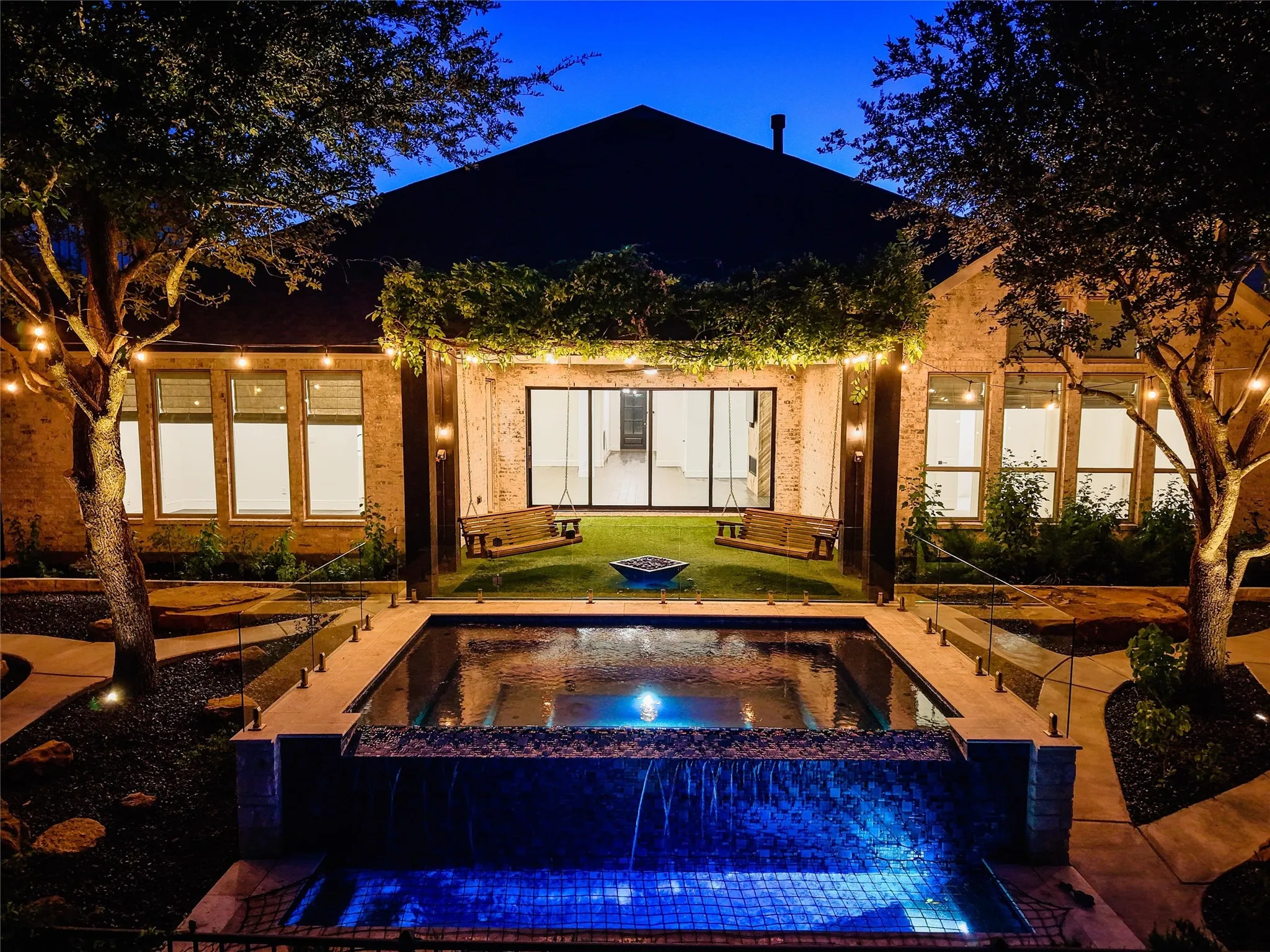 This closer twilight shot captures the heart of the outdoor oasis, where the glowing pool, lush pergola, and charming swing seating come together to create the ultimate setting for relaxing evenings.