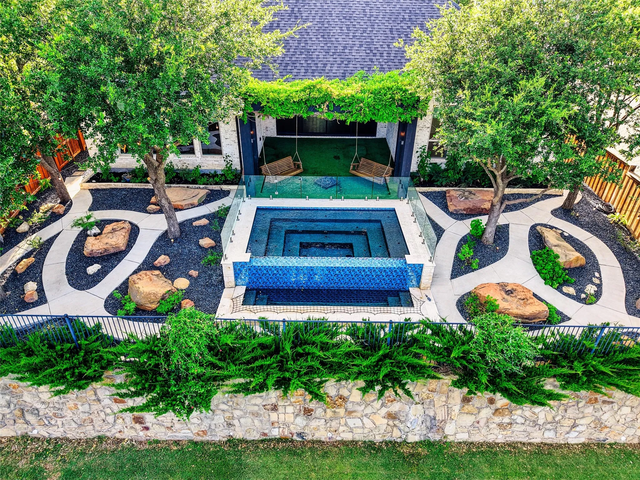 From above, the full vision comes to life—a dramatic resort-style pool, sculpted landscaping, winding concrete paths, natural boulders, mature trees, and a covered swing lounge hidden beneath a canopy of greenery.