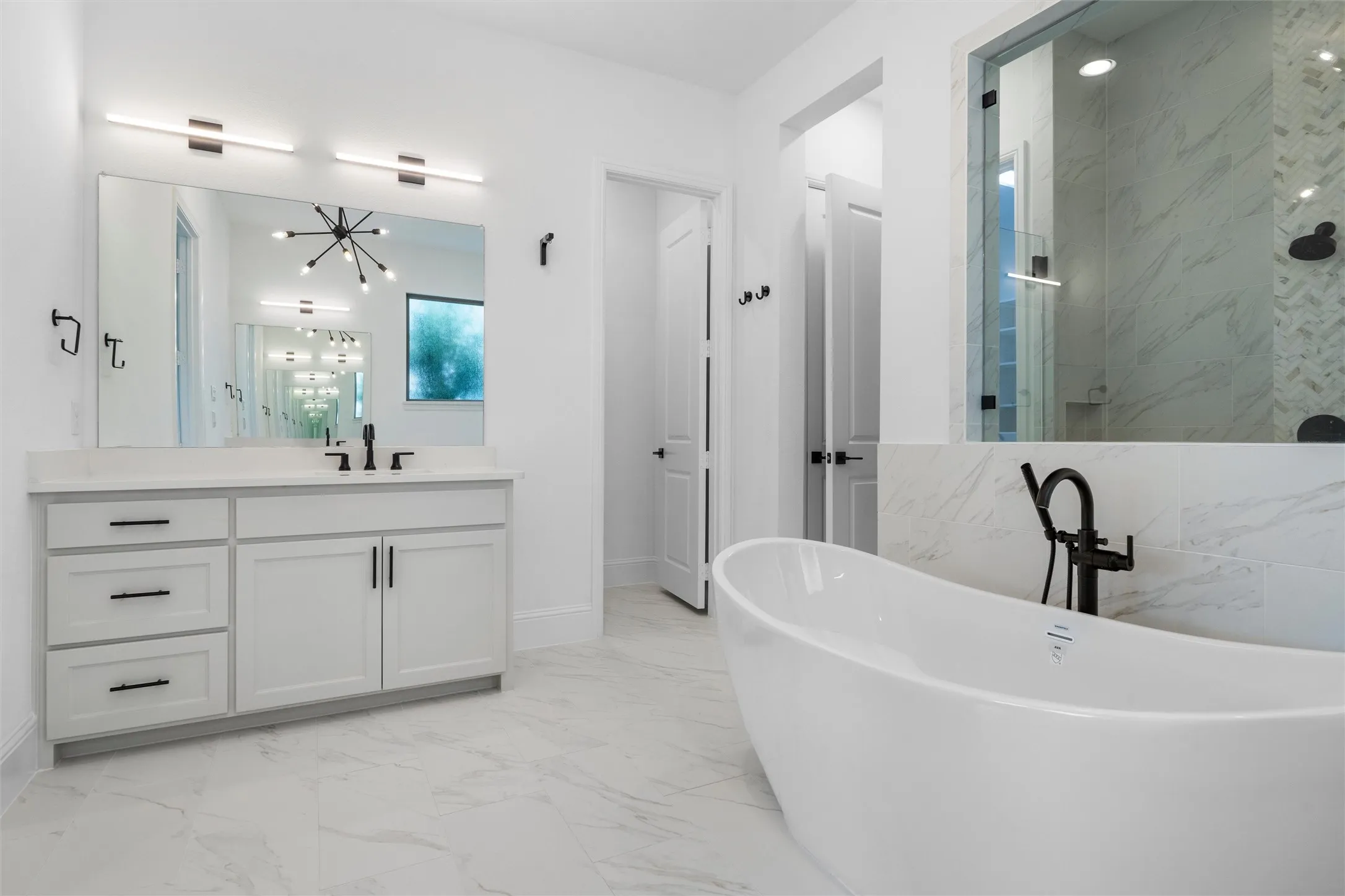 Modern finishes shine in this bright, refined bathroom with a spacious vanity, oversized mirror, designer lighting, freestanding tub, and a stunning marble-look shower enclosure.