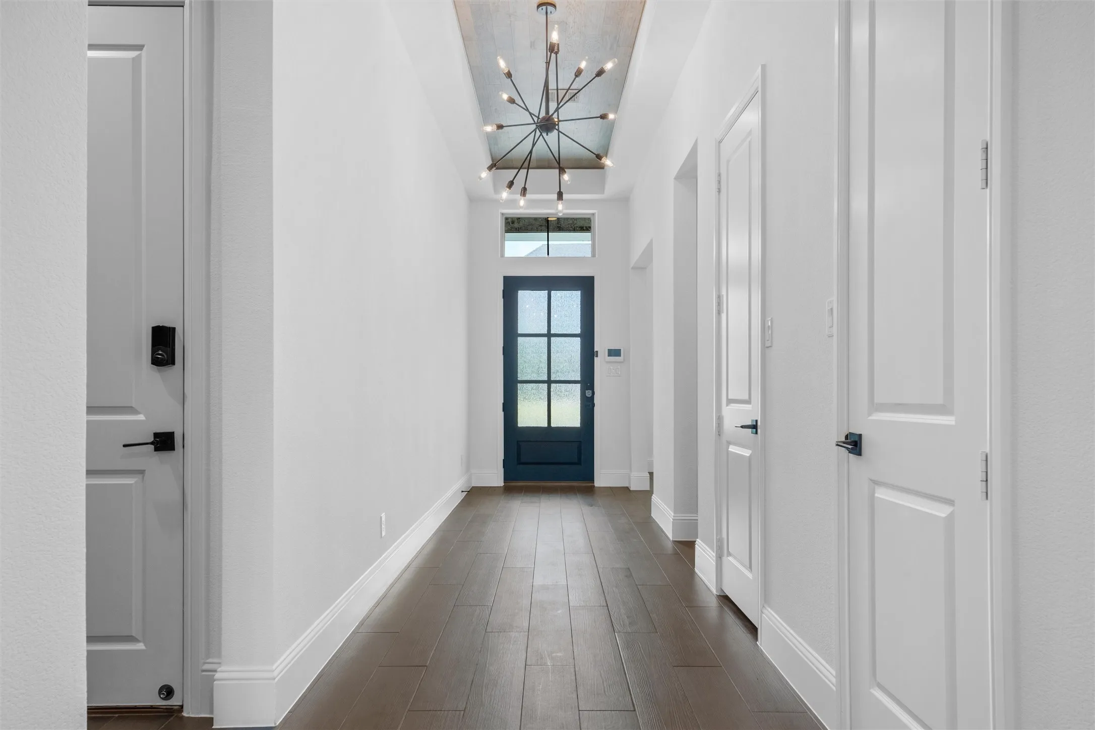 A dramatic entry hallway sets the tone with rich wood-look flooring, crisp white walls, a statement chandelier, and a bold black glass-paneled front door that delivers instant modern elegance.