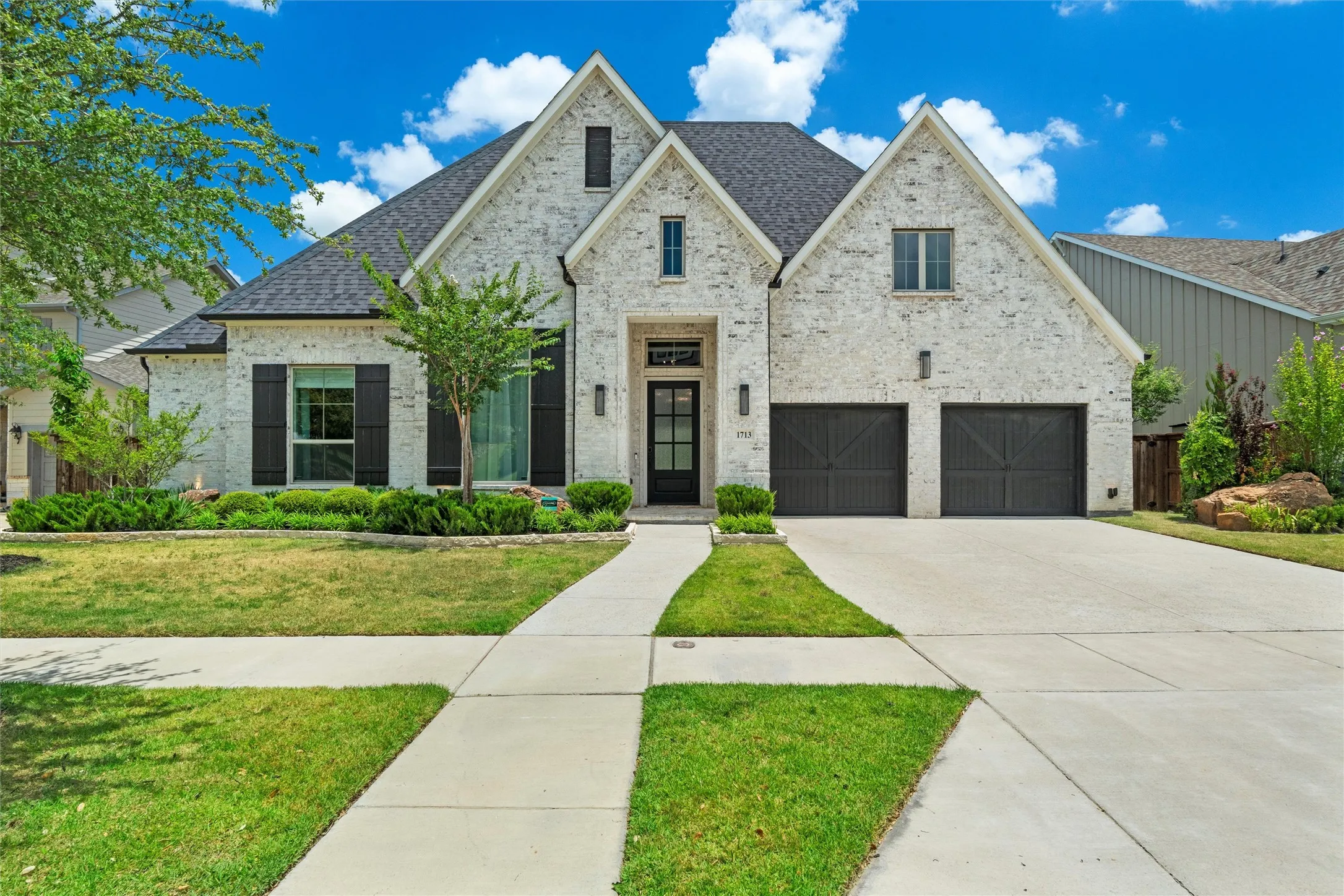 A bold first impression starts here—crisp white brick, soaring gables, black designer shutters, and dramatic carriage-style garage doors create a front elevation that feels straight out of a luxury home magazine. Framed by manicured landscaping and a vivid Texas sky, this home does not just have curb appeal—it owns the street.