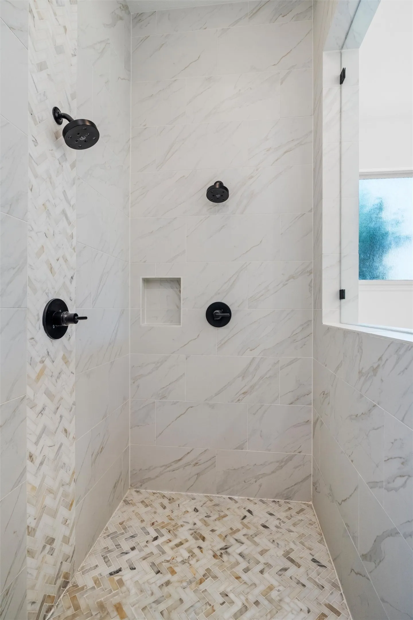 The walk-in shower is a true statement piece, featuring floor-to-ceiling marble-look tile, a herringbone mosaic accent wall and floor, matte black fixtures, dual showerheads, and a built-in niche.
